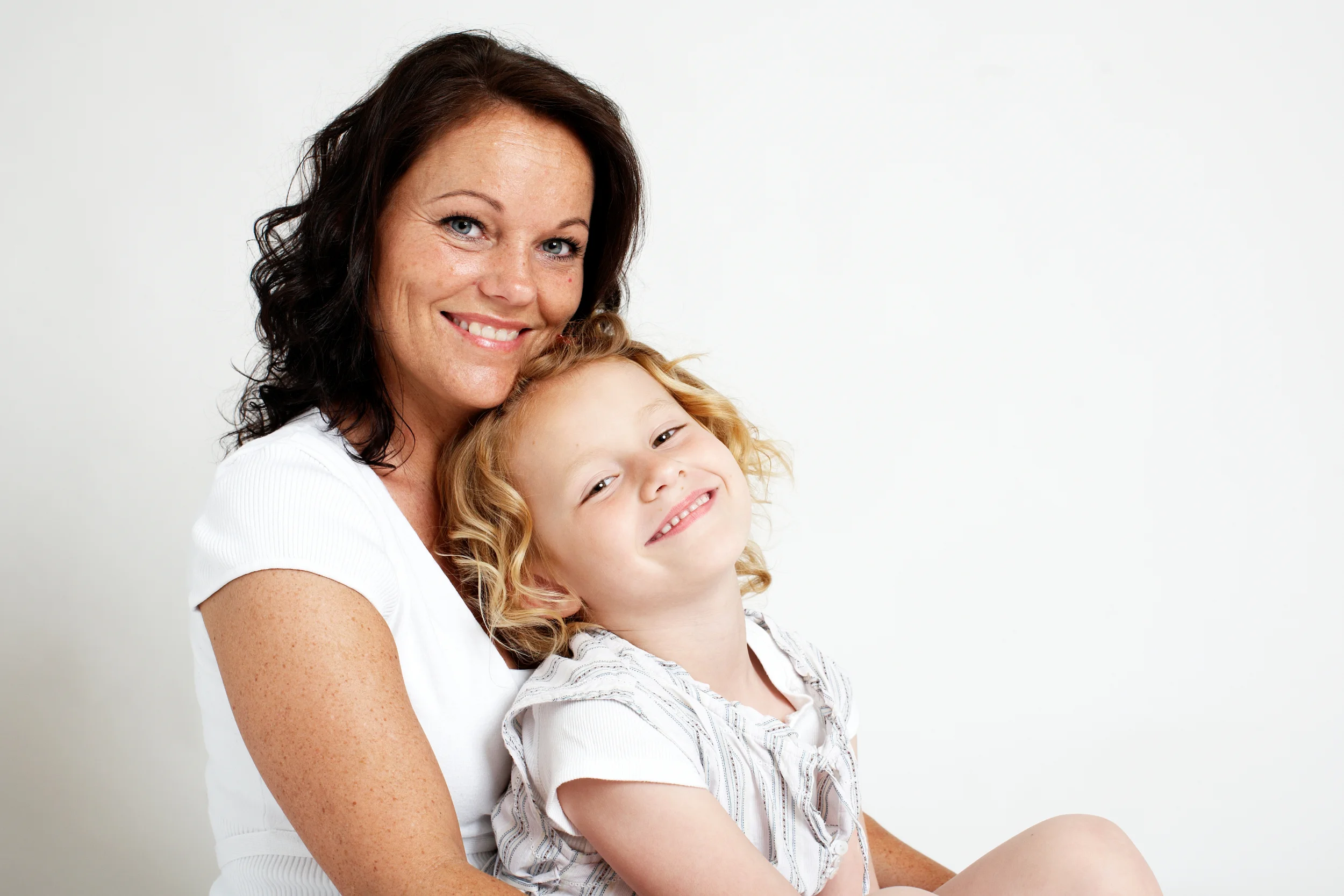 Familieportræt af smilende mor med mørkt hår, der krammer sin lille datter med krøllet lyst hår. Hjertevarm stemning.