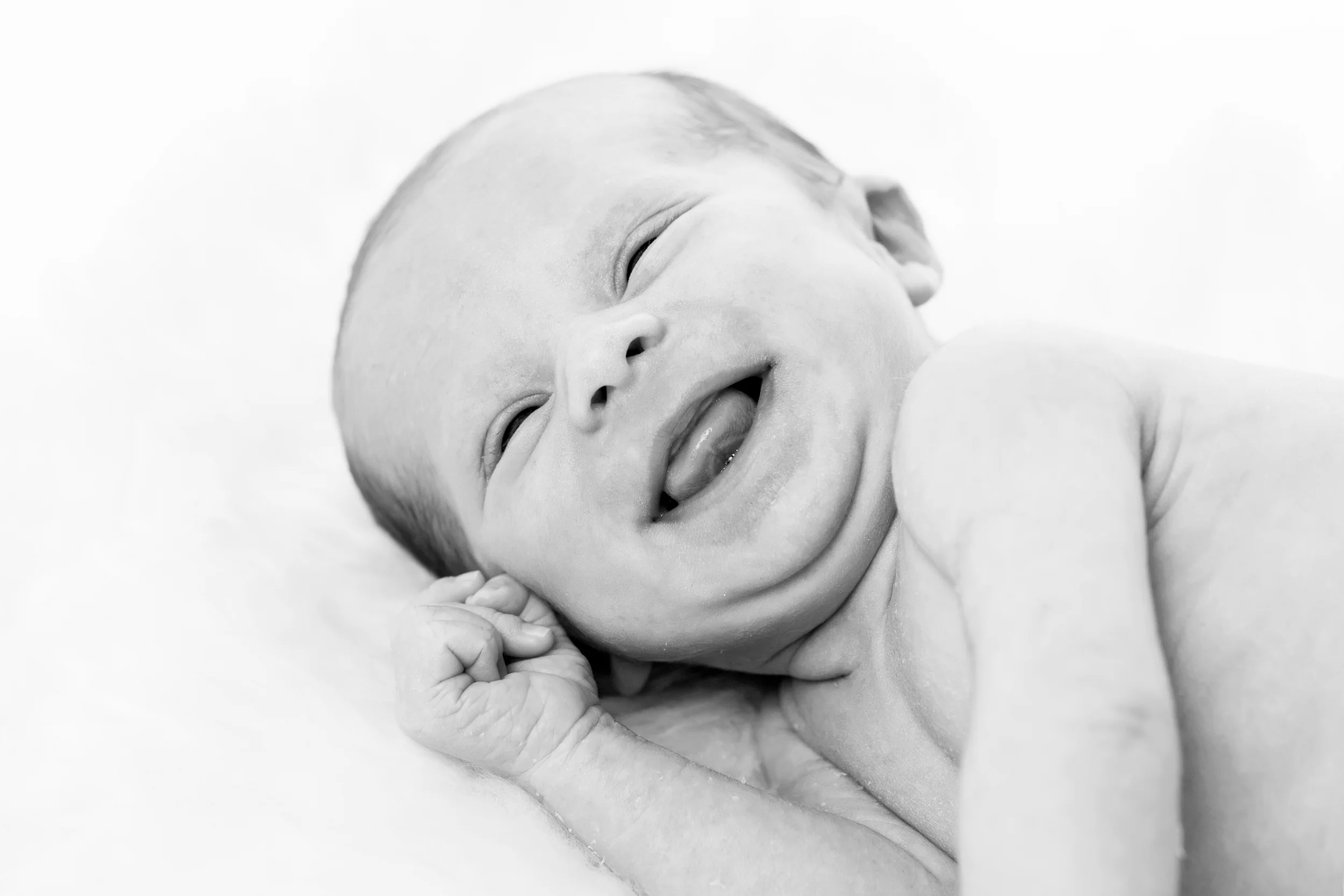 Baby fotografering af glad nyfødt, der smiler bredt med tungen fremme, i blødt sort-hvidt lys.