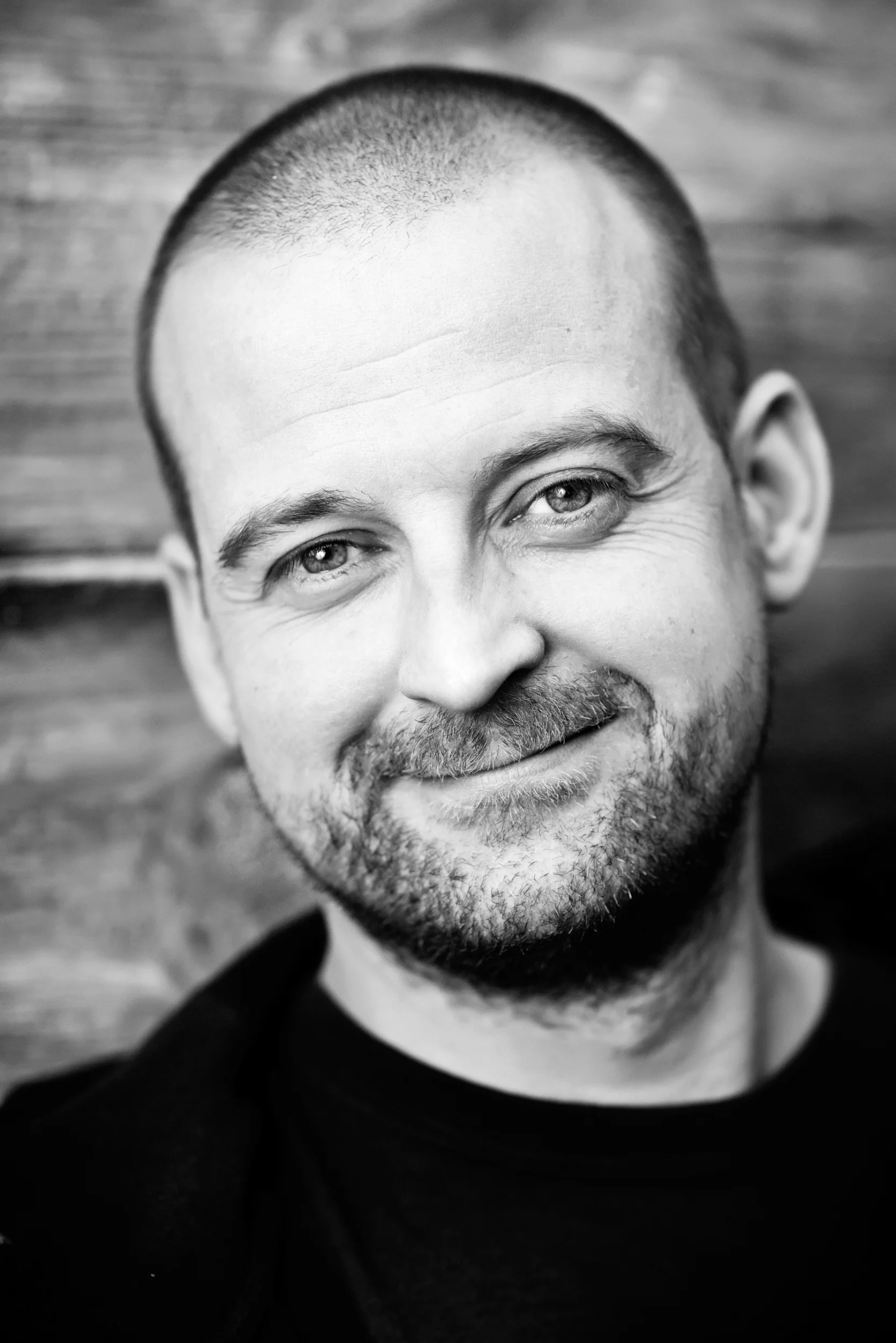 Sort/hvid portrætfoto af smilende mand med kortkronet skæg og lyst, nyklippet hår, i blød belysning.