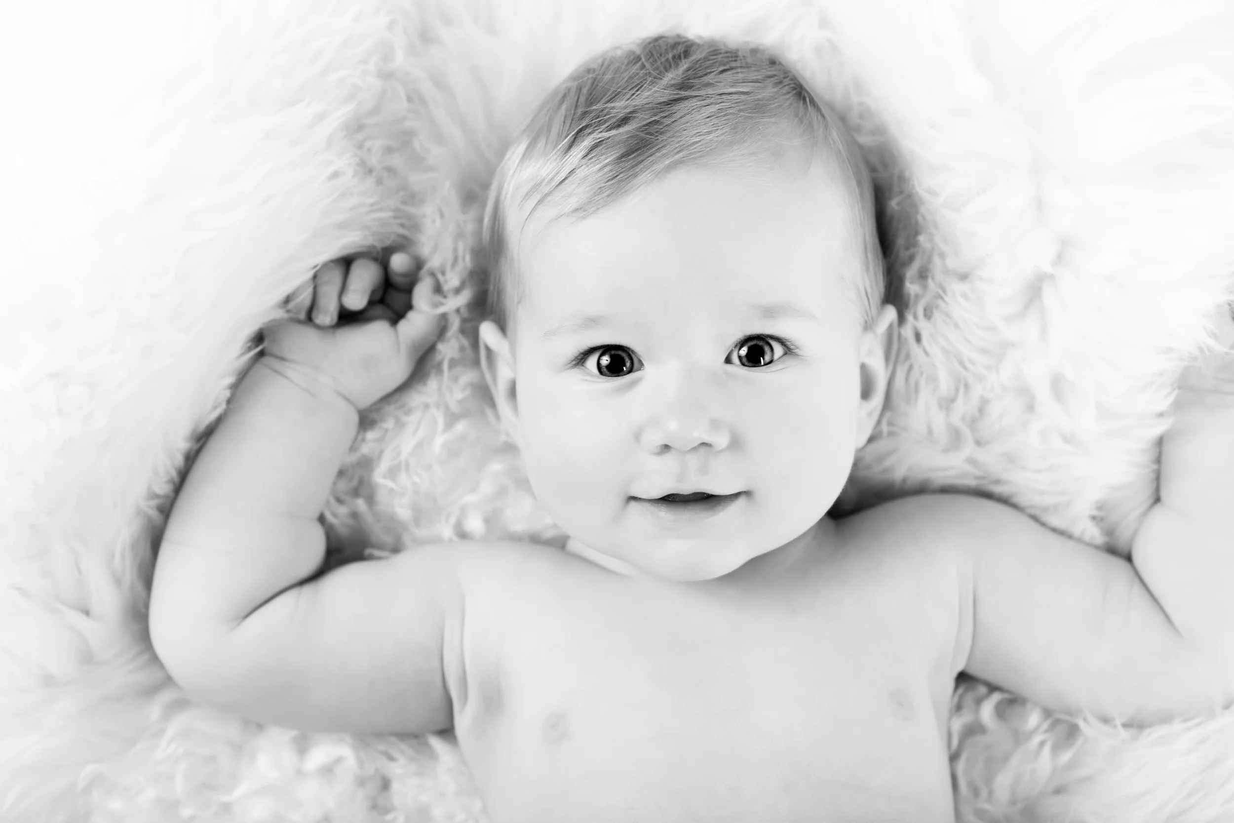Nærbillede af glad baby med store øjne i sort/hvid, liggende på et blødt, hvidt tæppe. Et skønt babyfoto.