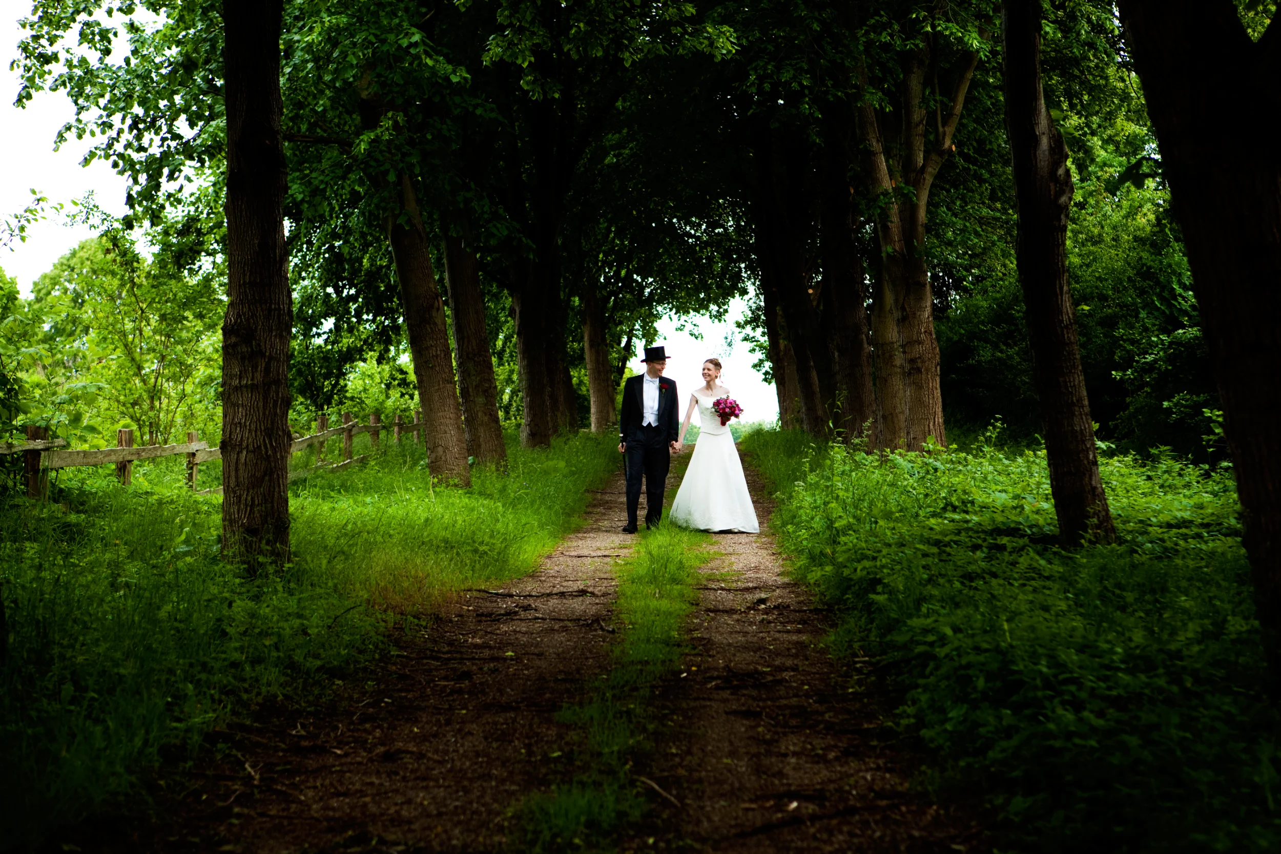 Bryllupsfoto: Et brudepar går hånd i hånd på en grussti flankeret af grønne træer og frodig vegetation i smukt, blødt lys.
