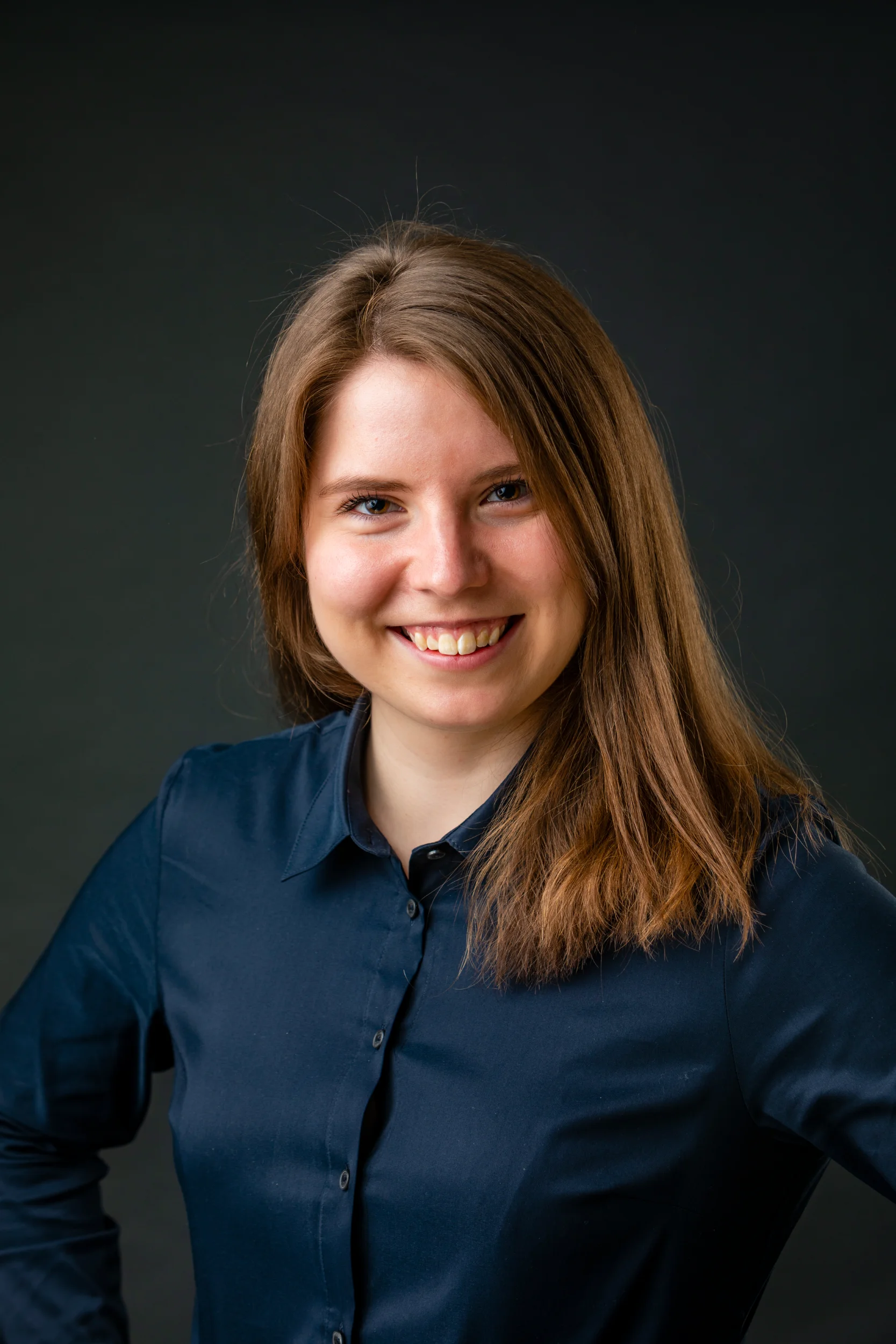 Portrætfoto af smilende kvinde med brunt hår i mørkeblå skjorte mod mørk baggrund, et autentisk portræt.