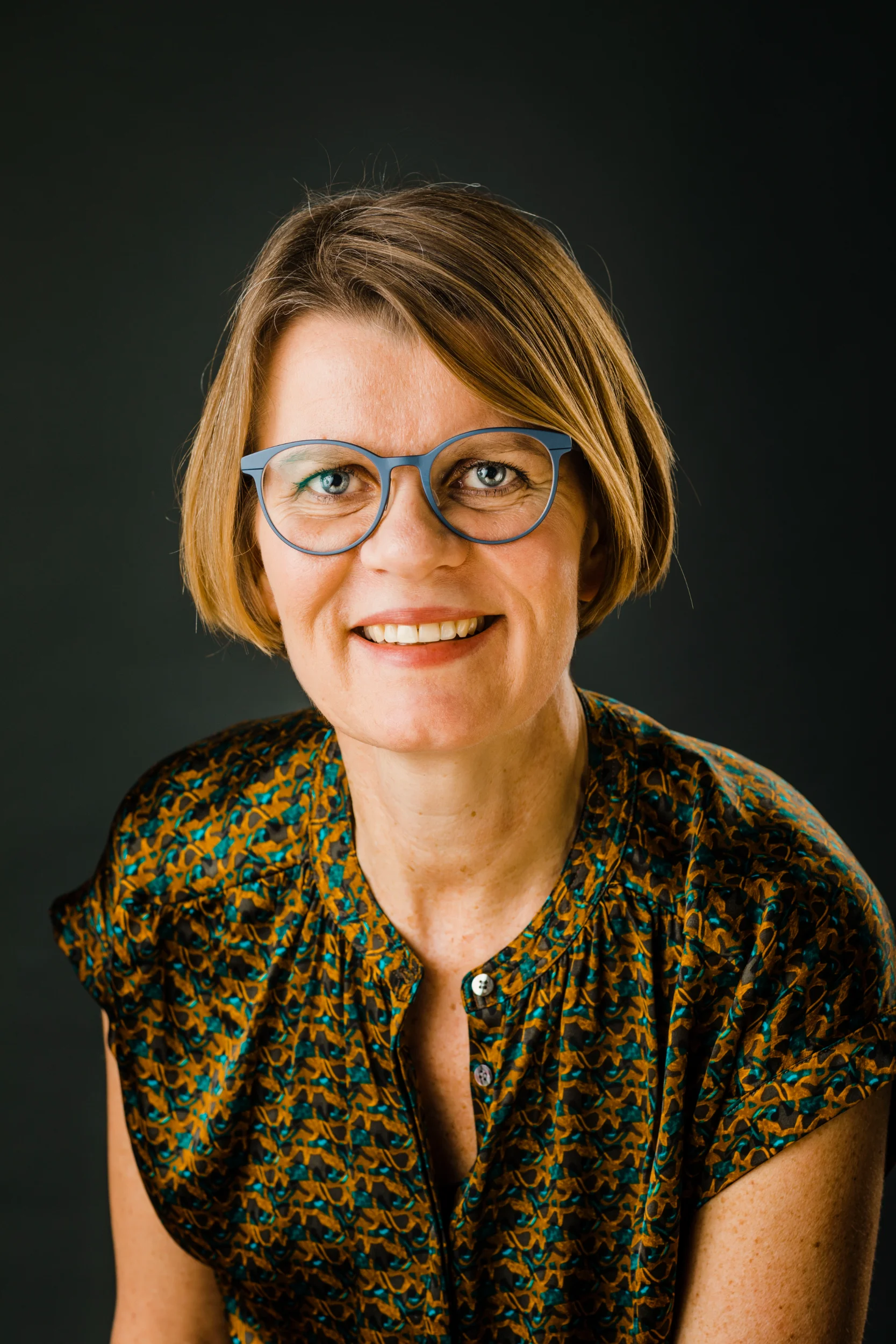 Portrætfoto af smilende kvinde med kort, lyst hår og blå briller, lys sat mod mørk baggrund.
