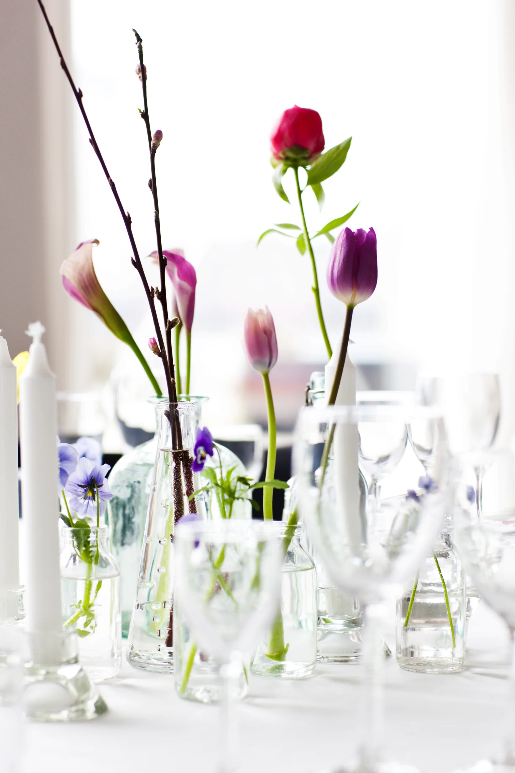 Festlig bryllupsborddekoration: Smukke blomster som lilla tulipaner og lyserøde kallaer i klare vaser på hvid dug.