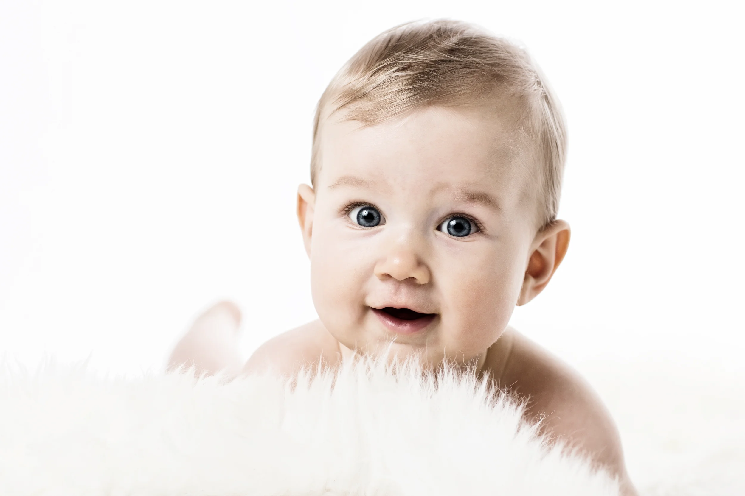 Glad baby med blå øjne og lyst hår ligger på maven, smilende på et hvidt lammefæld i blødt studielys. Babyfotografering.