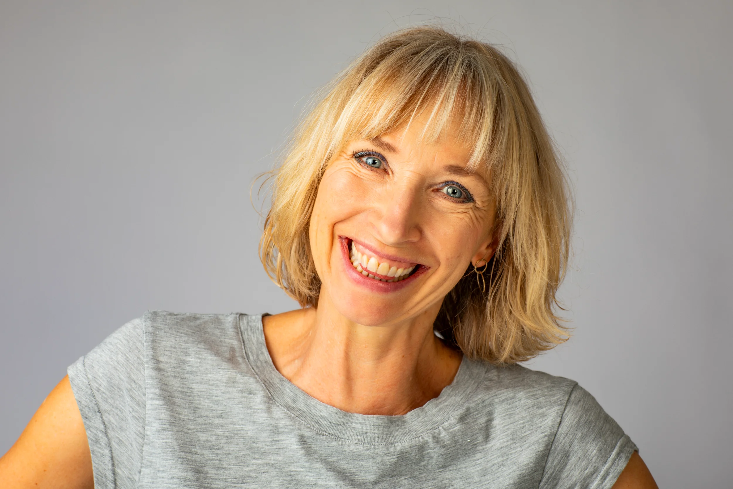 Portrætfoto af smilende blond kvinde med glade blå øjne i naturligt varmt lys mod en grå baggrund.