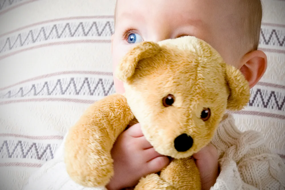 Nærbillede af baby, der holder en lille bamse op til ansigtet med store, blå øjne kiggende frem.