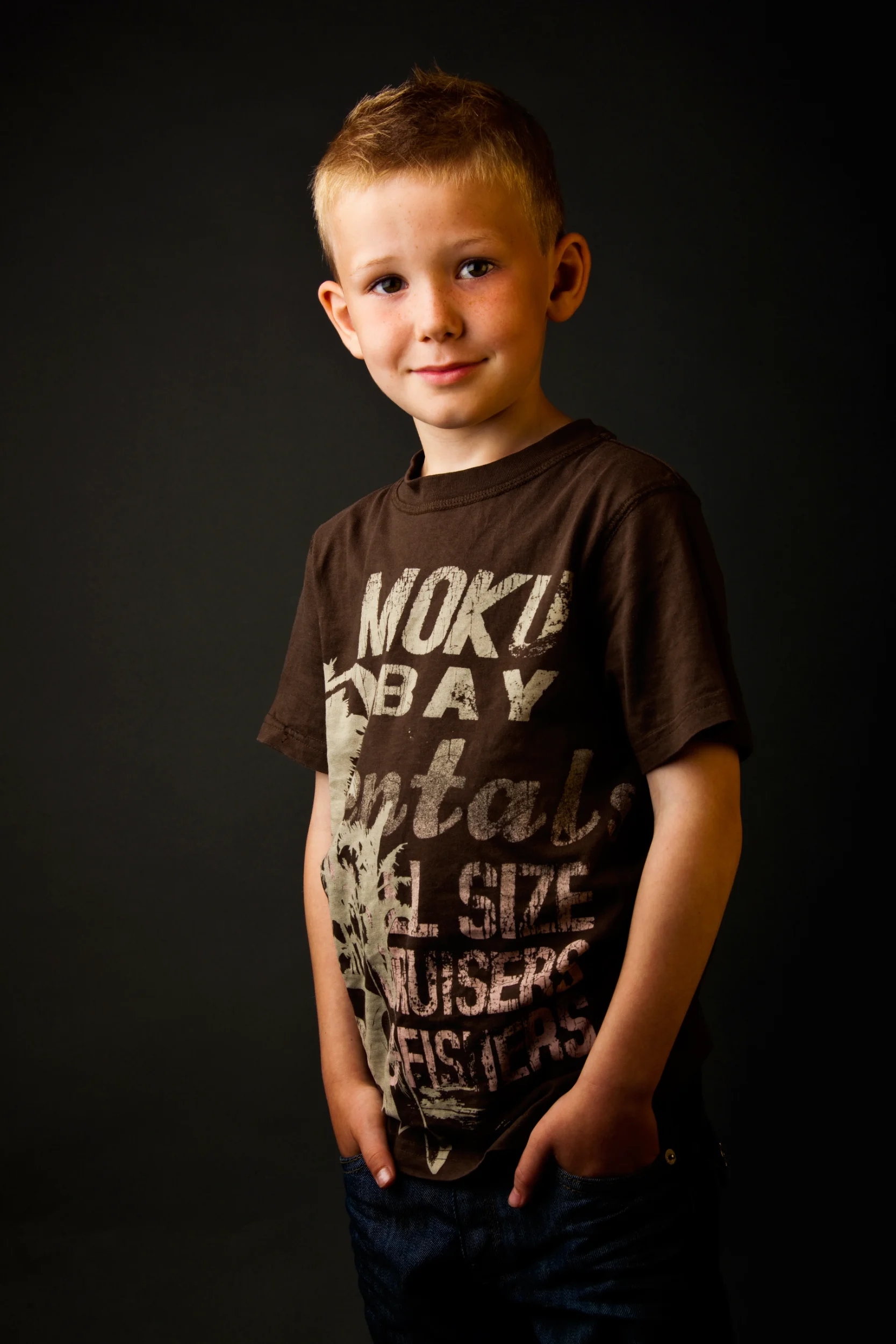 Portrætfoto af glad, blond skoledreng med hænderne i lommen, iført brun t-shirt, mod mørk baggrund med blødt lys.