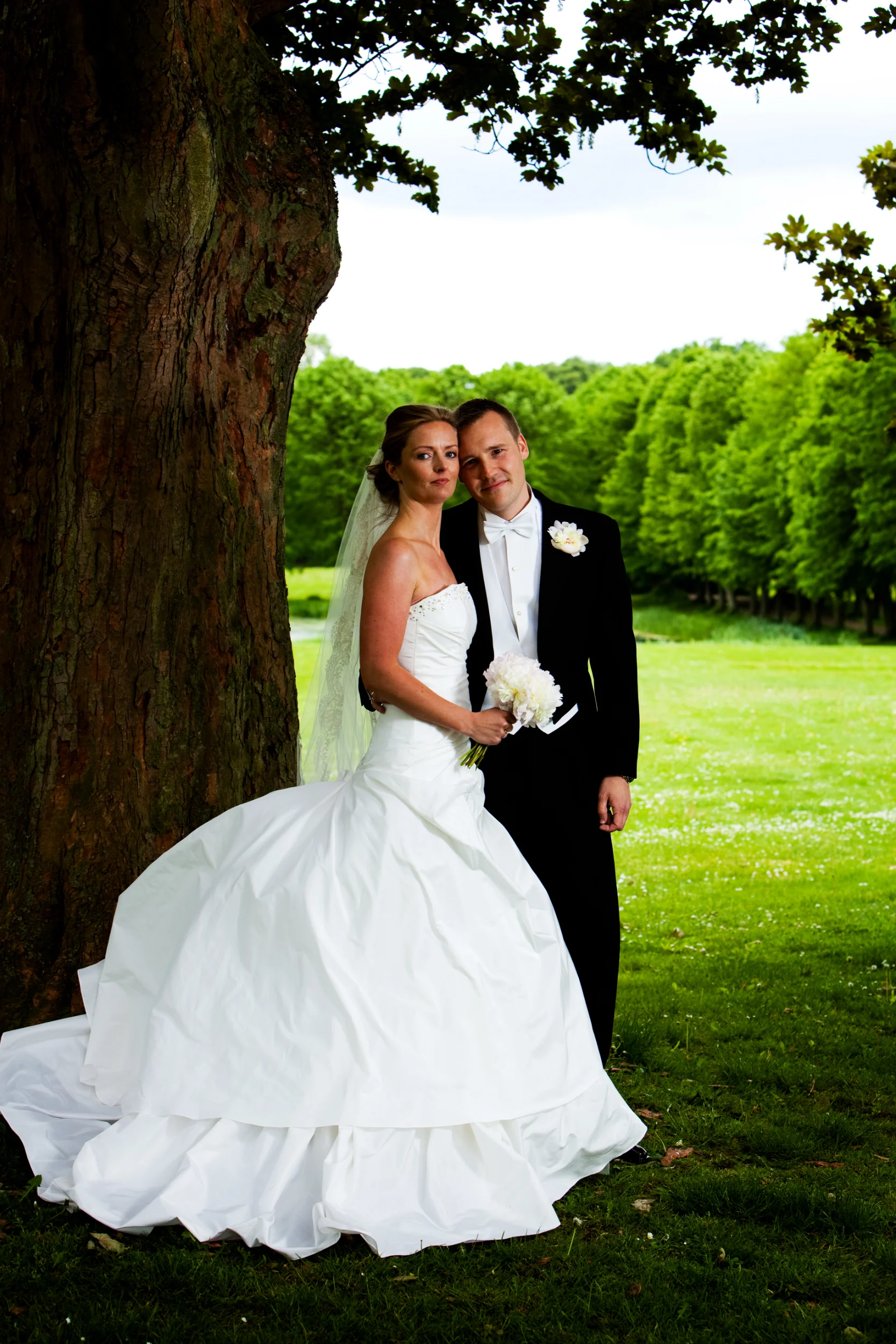 Bryllupsfoto af brudepar under et stort træ i blødt lys, omgivet af græs og grønne træer i baggrunden.