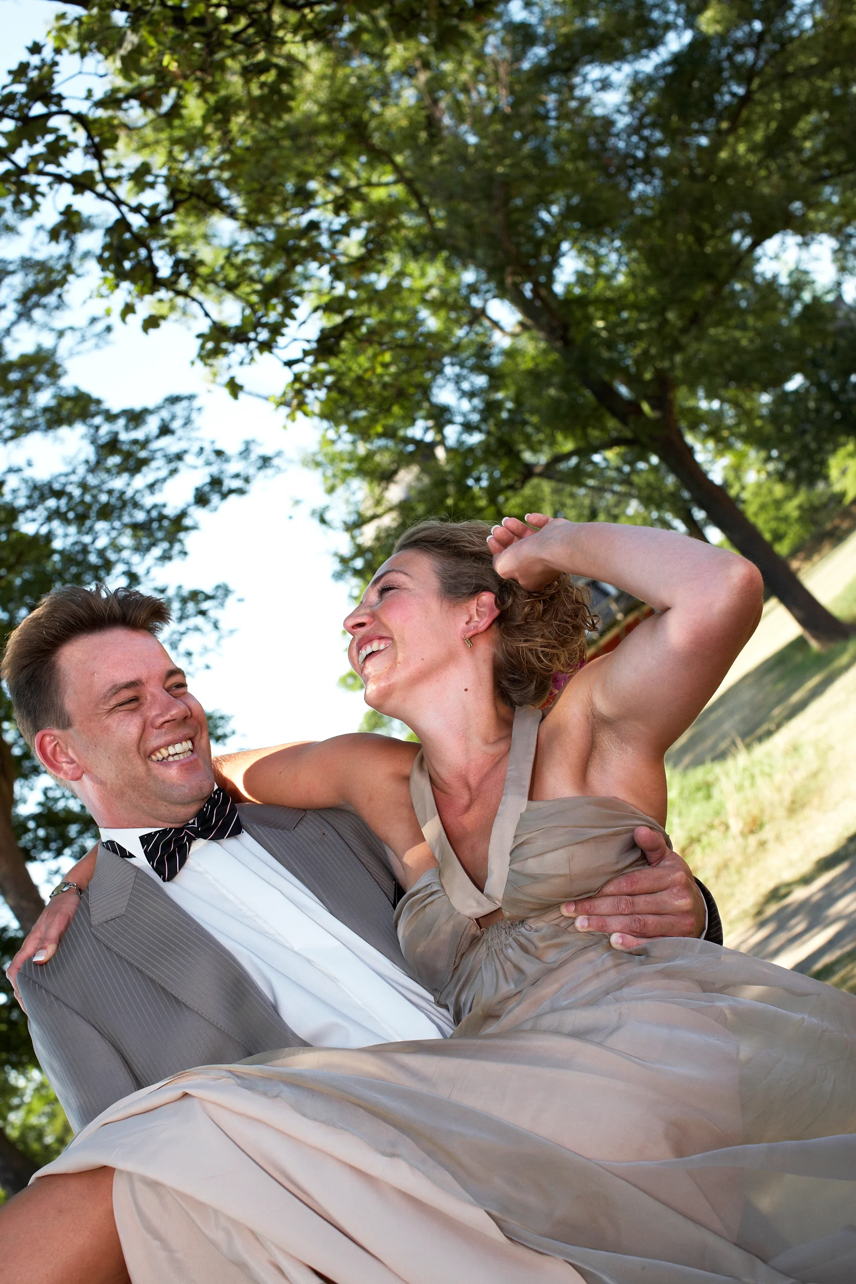 Bryllupsfoto af smilende brudepar i naturlige omgivelser, hvor manden løfter kvinden og de griner sammen. Fokus på ægte gl...