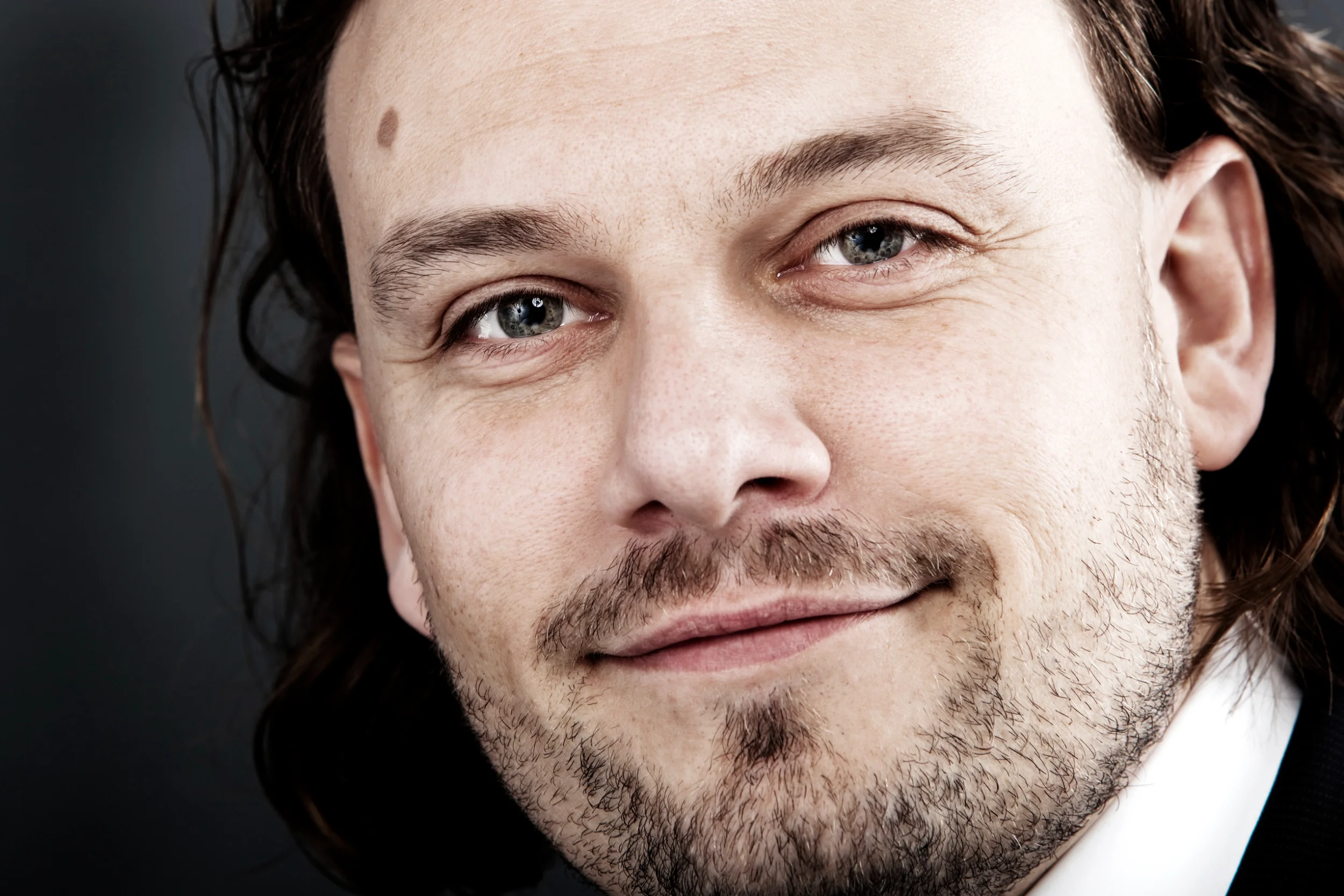 Nært portrætfoto af en smilende mand med skæg og langt mørkt hår, der kigger ind i kameraet med venlige øjne.