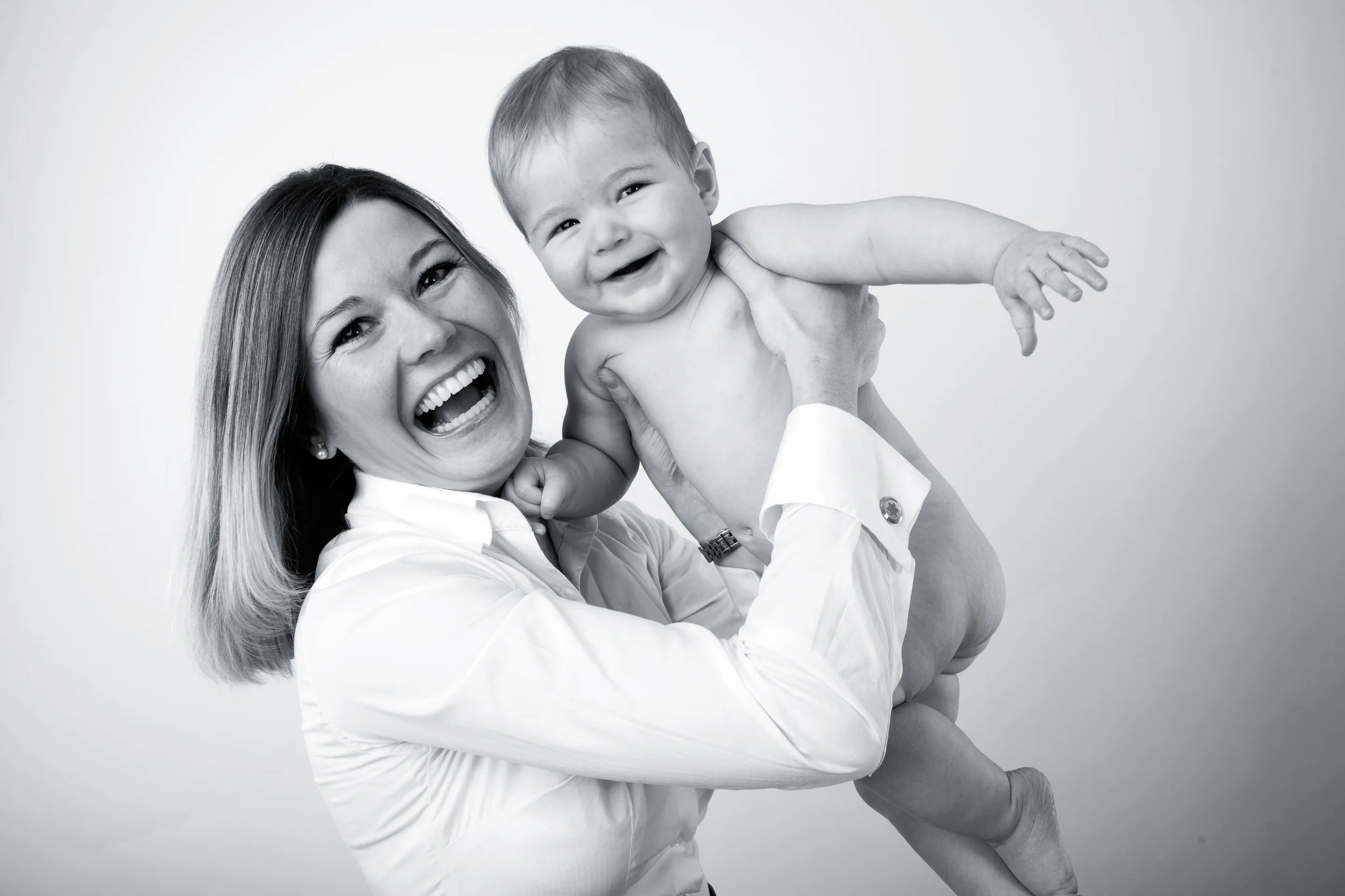 Glad mor løfter sin leende baby op i luften, tæt på i sort/hvid med skarp belysning. Et dejligt babybillede.