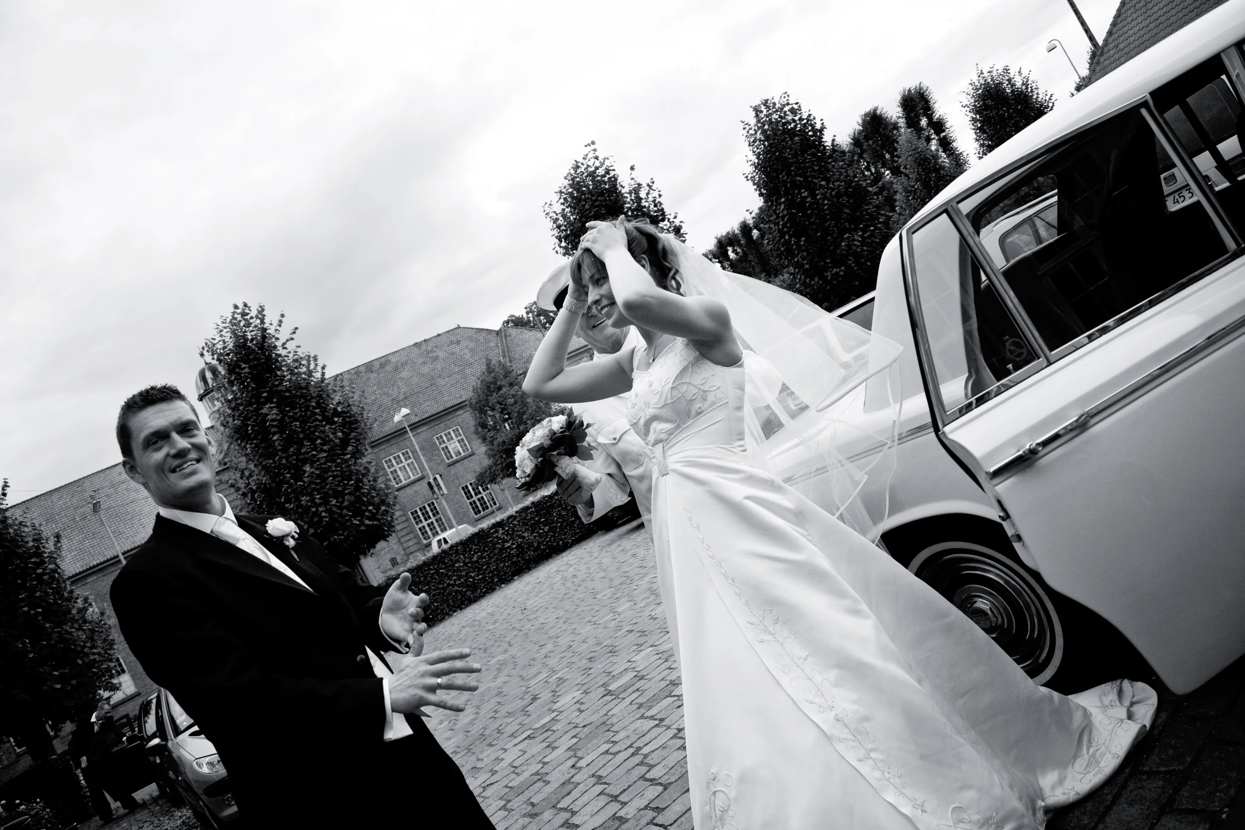 Bryllupsfoto i s/h af brudeparret der netop er ankommet i bil og griner, med bruden der holder sløret på plads.