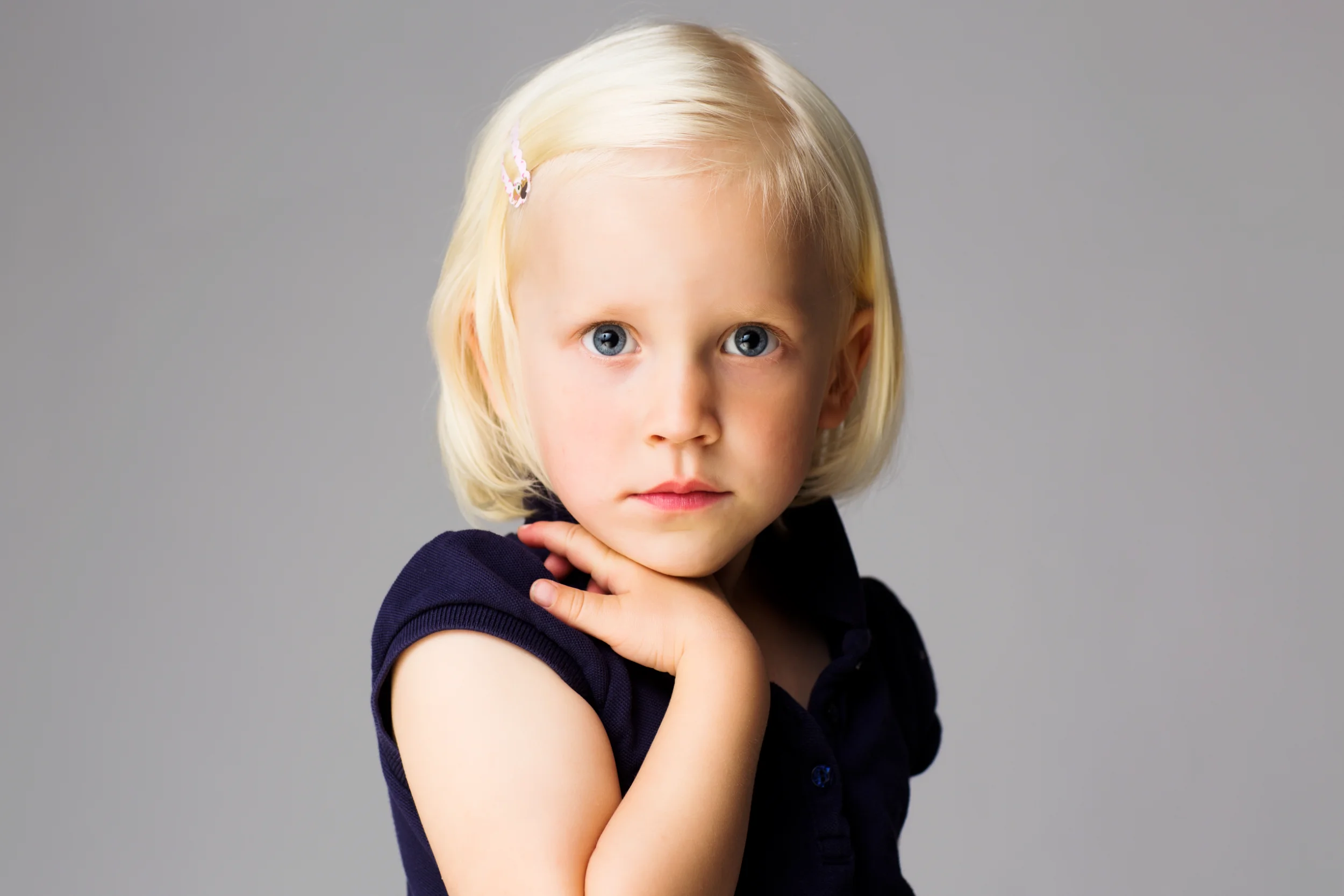 Portrætfoto af lille pige med lyst, kort hår og blå øjne, der hviler hånden ved halsen med grå baggrund.