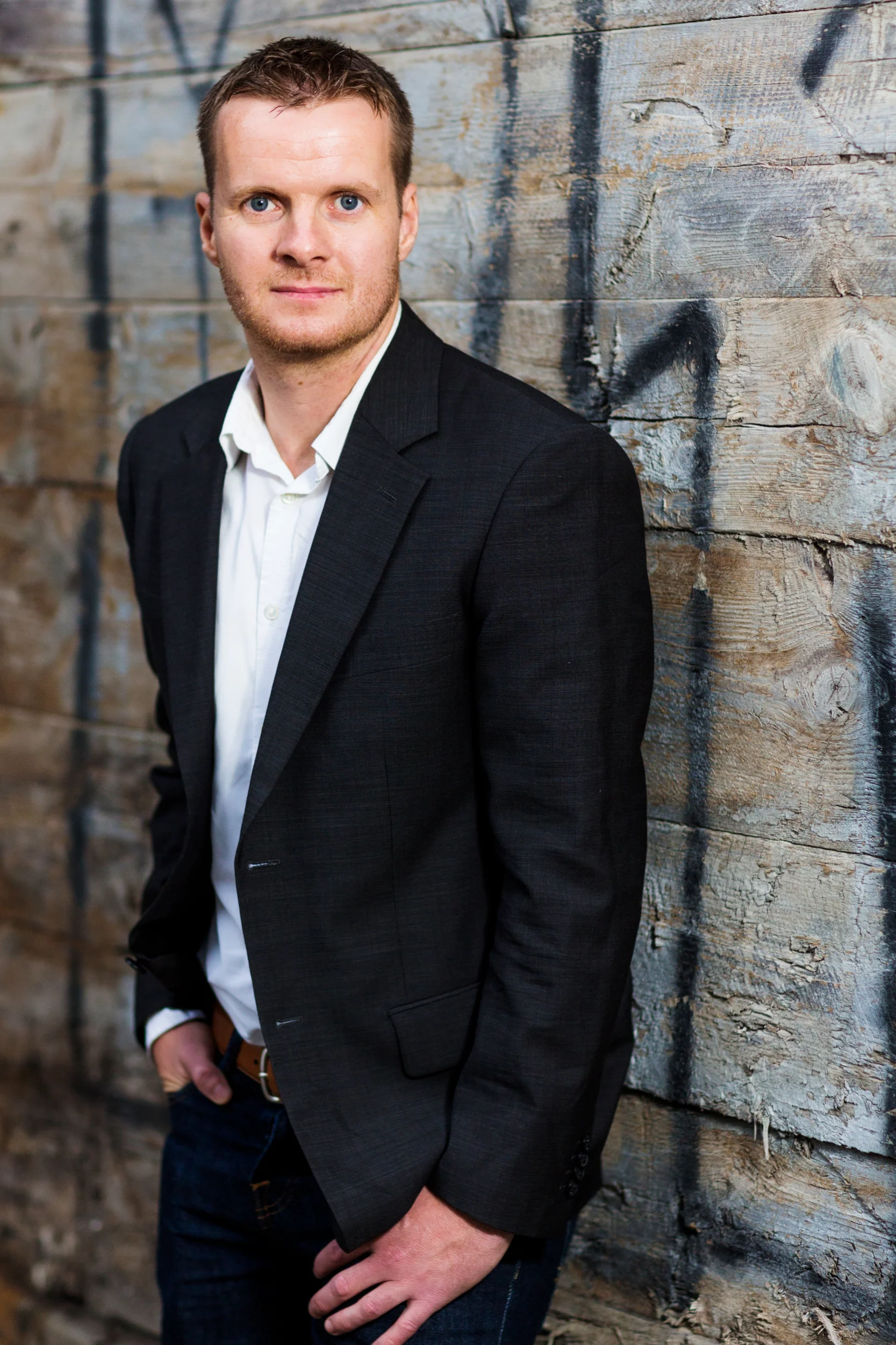 Portrætfoto af mand i mørkt jakkesæt og hvid skjorte, med blåt blik, stående mod rå trævæg med graffiti. Ser selvsikker ud.