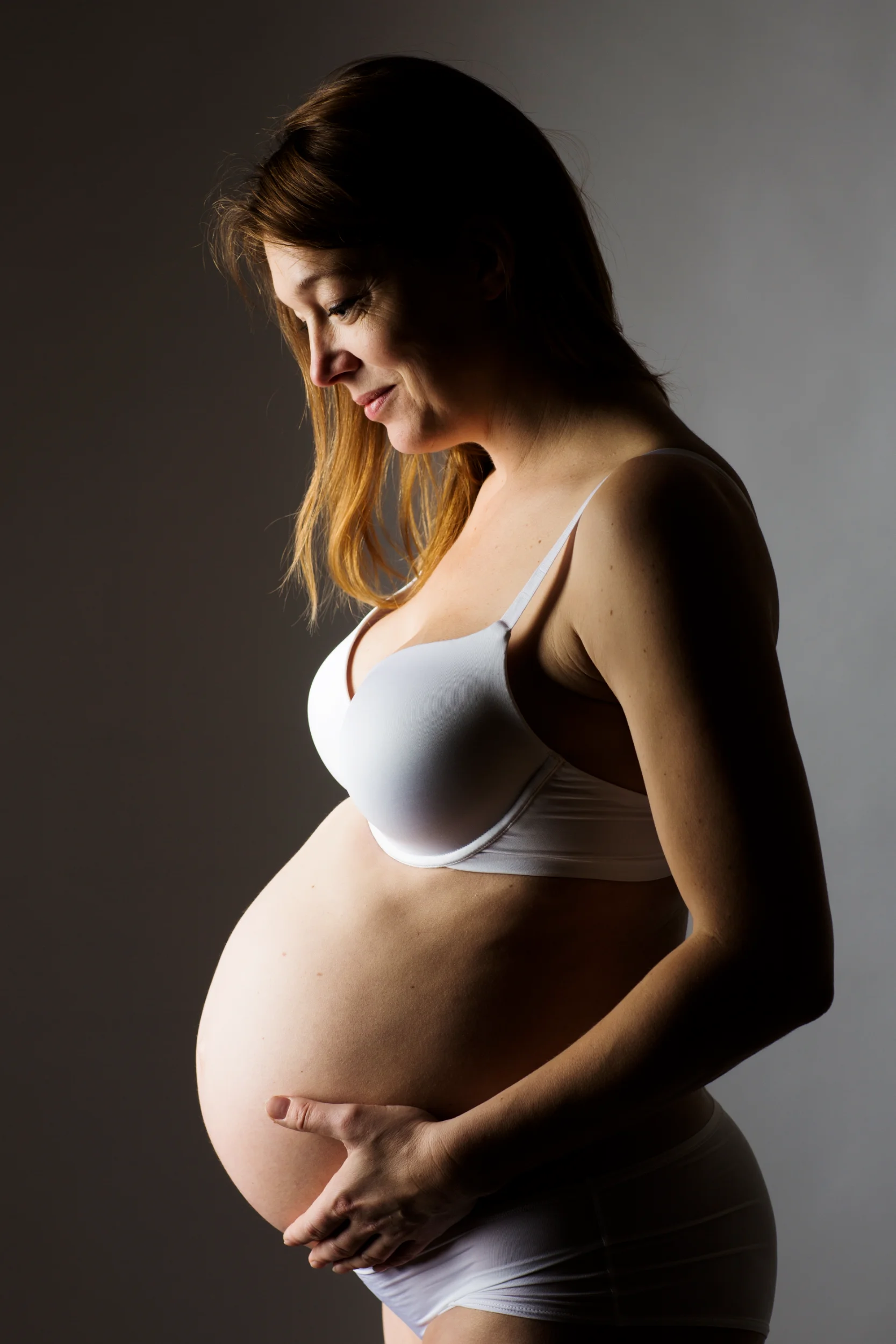 Gravid kvinde fotograferet i profil, iført hvidt undertøj, holder om sin mave i blødt sidelys. Graviditetsfotografering.