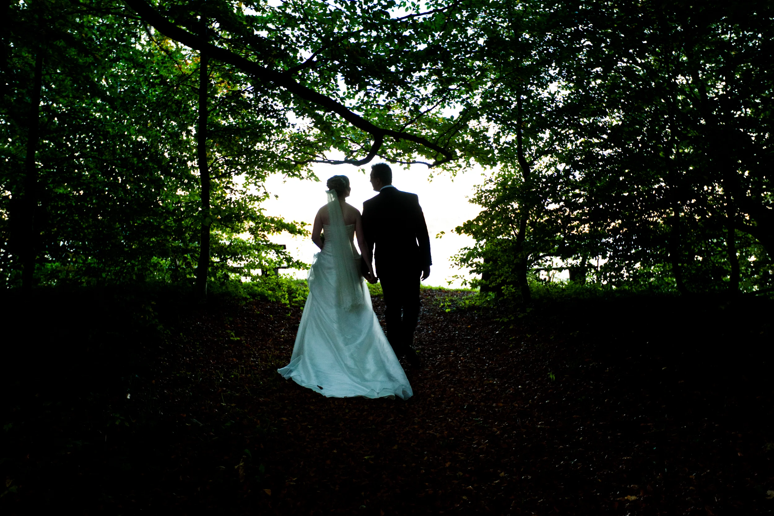 Bryllupsfoto af brudepar i silhuet, der går hånd i hånd ad skovsti med lys for enden. Stemningsfuld fotografering.