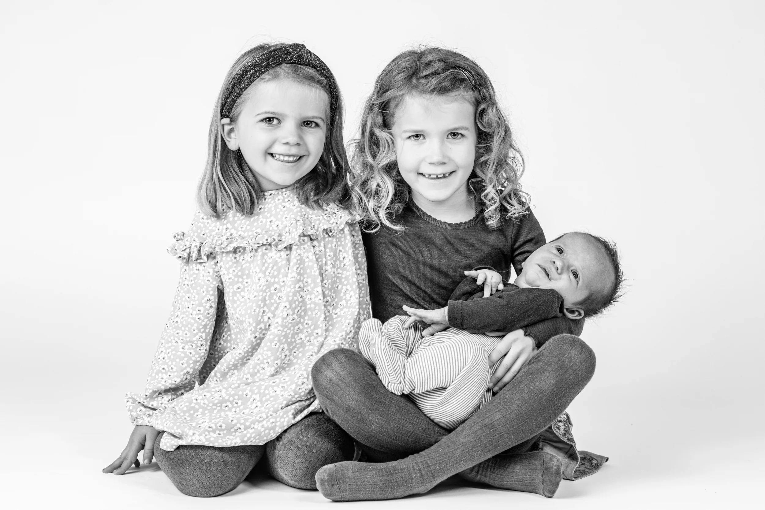 Babyfotografering med to søde piger og deres nyfødte søskende smilende mod kameraet i blødt lys.