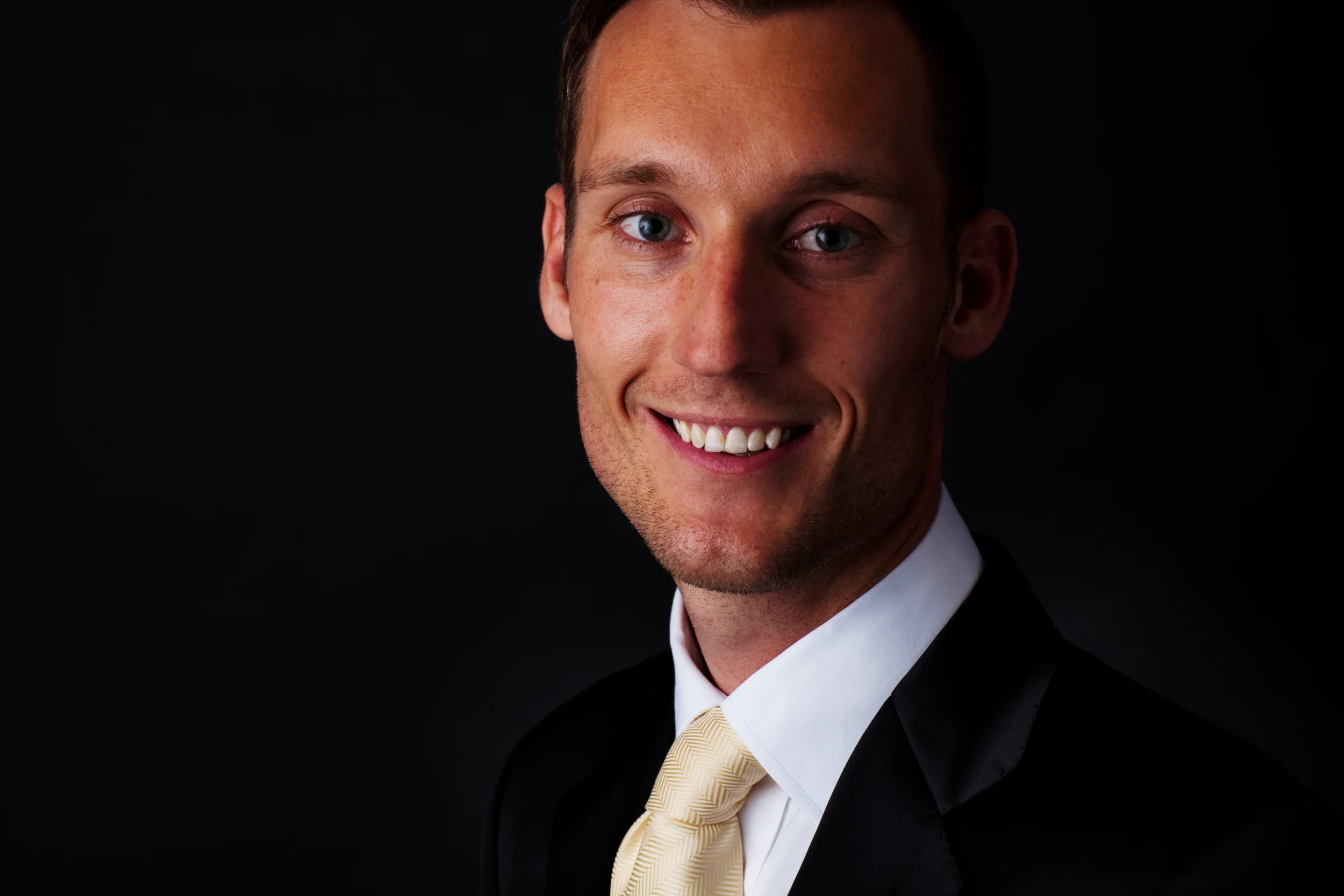 Professionelt portrætfoto af smilende mand i sort jakkesæt med guldbind, belyst fra siden mod mørk baggrund.