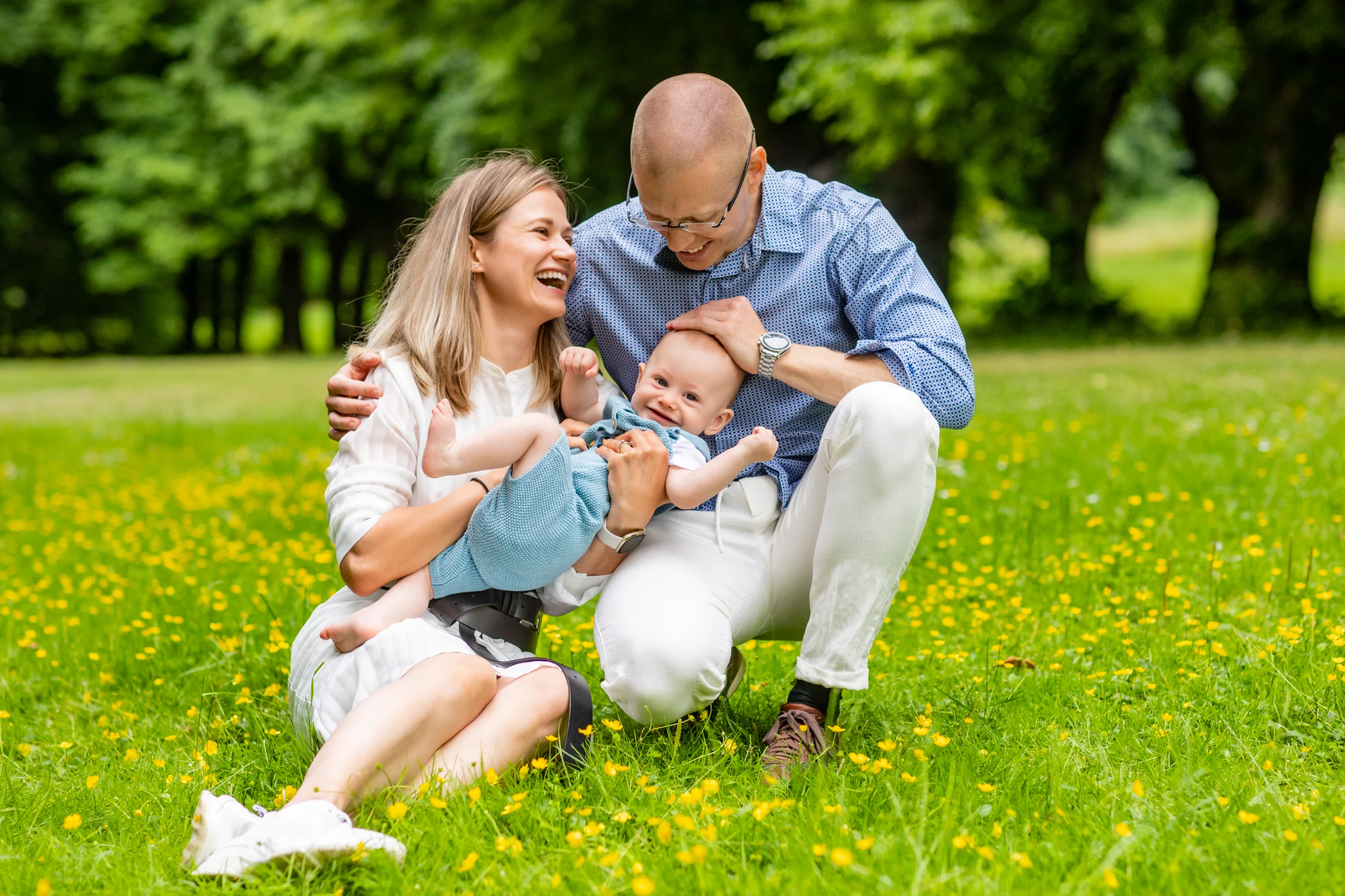 Familie fotografering i Virum. Smilende forældre hygger om glad baby i armene på en grøn sommereng med gule blomster.