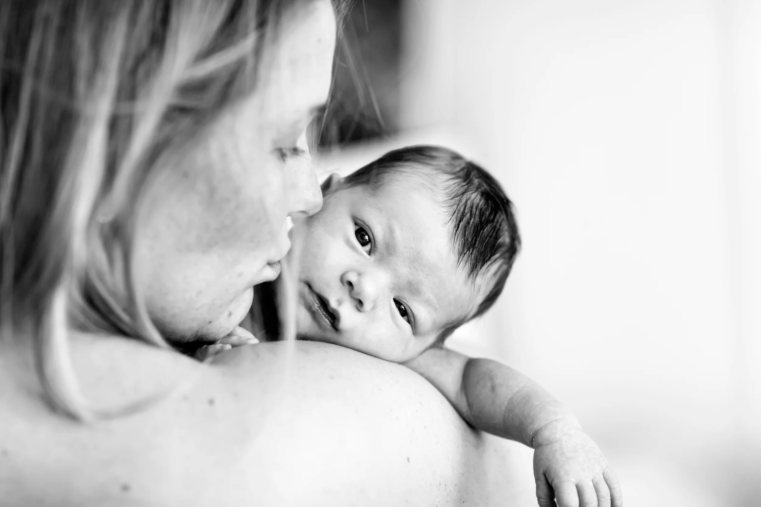 Sort/hvidt babyportræt: En lille baby kigger direkte ind i kameraet fra mors trygge skulder i blødt lys.
