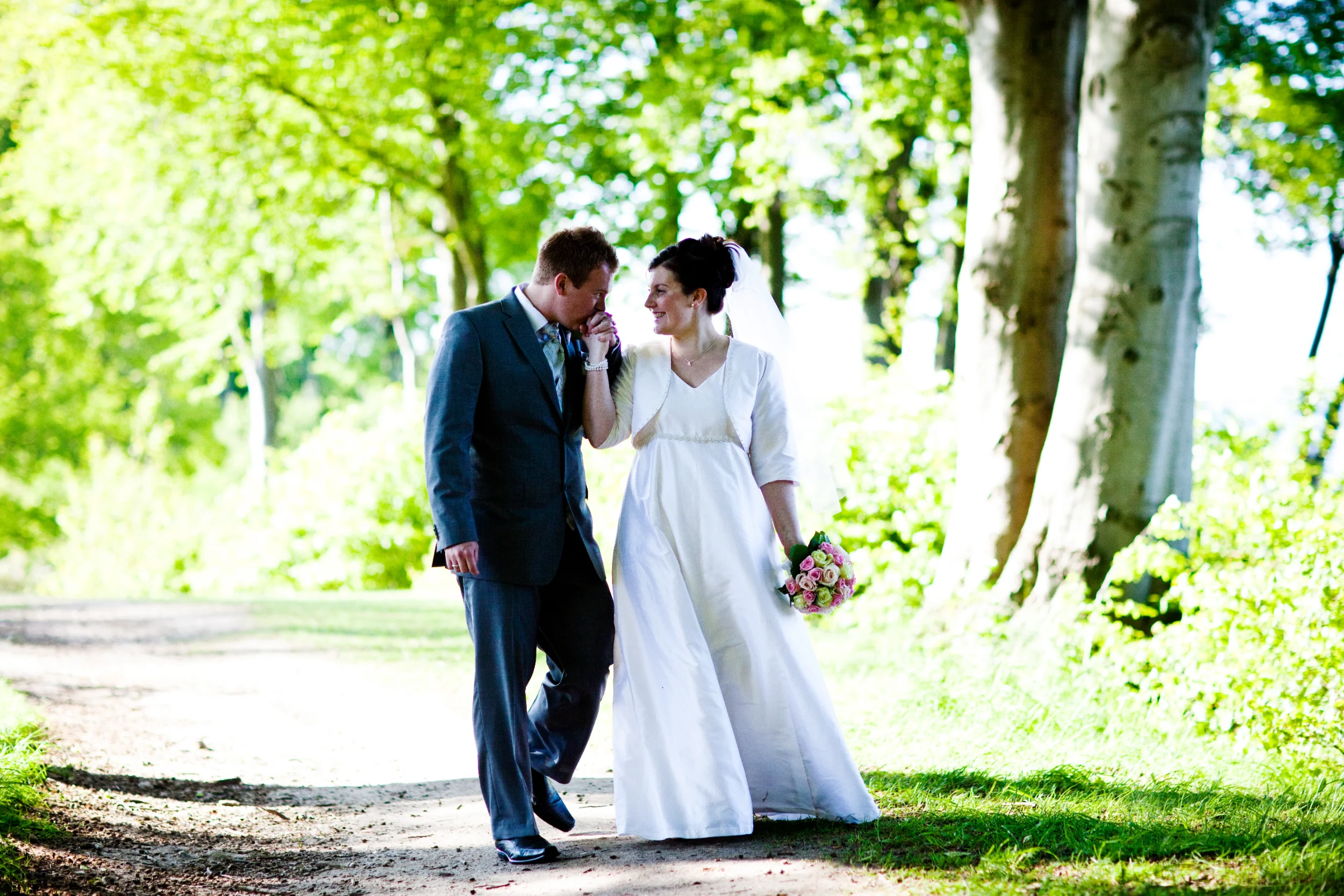 Bryllupsbillede af smilende par i sort-hvid. Manden kysser kvindens hånd, de går på sti i skov med blødt lys.