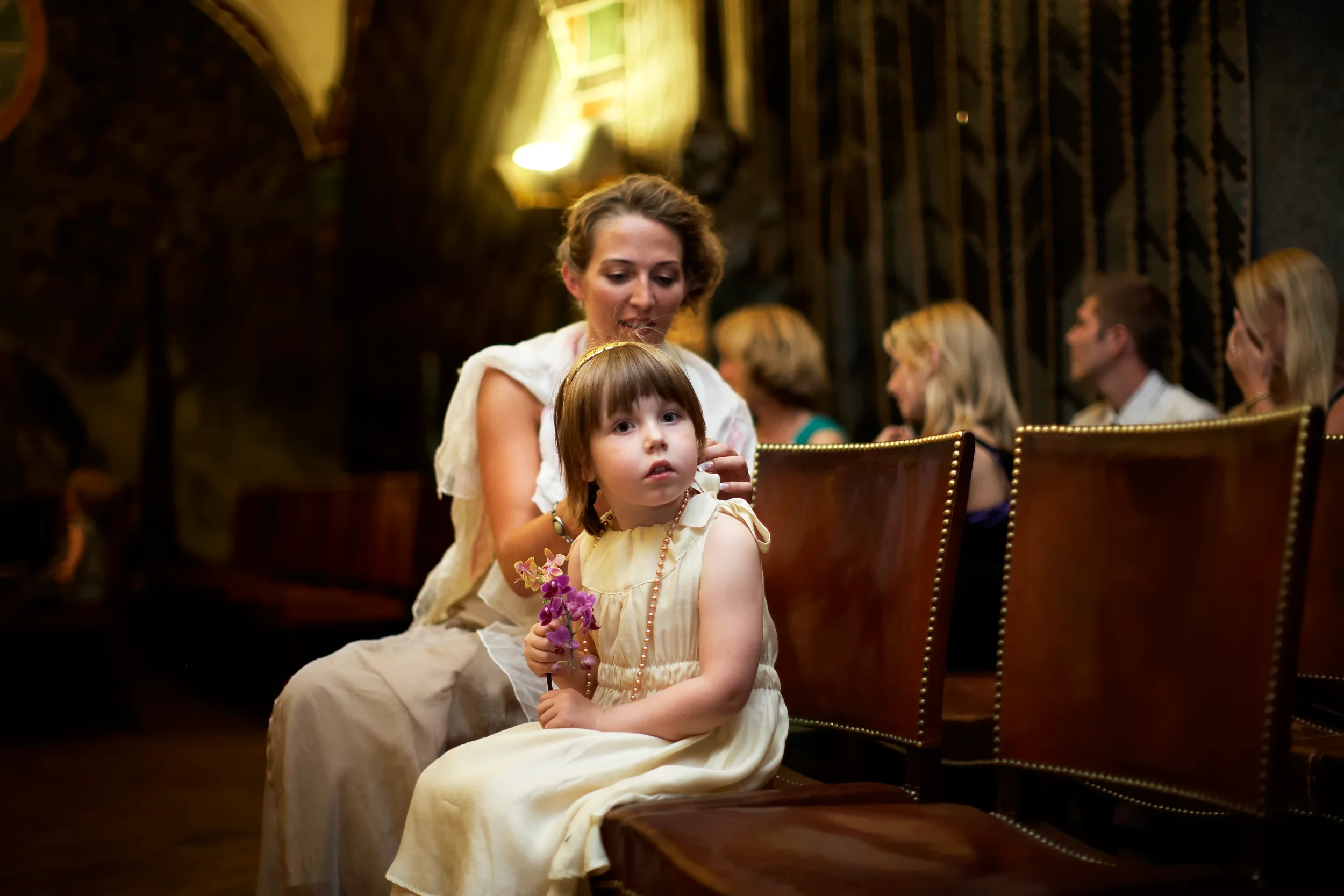 Bryllupsfotografi af mor, der kærligt ordner sit barns hår; den lille pige sidder med blomster og perlekæde i varmt lys.