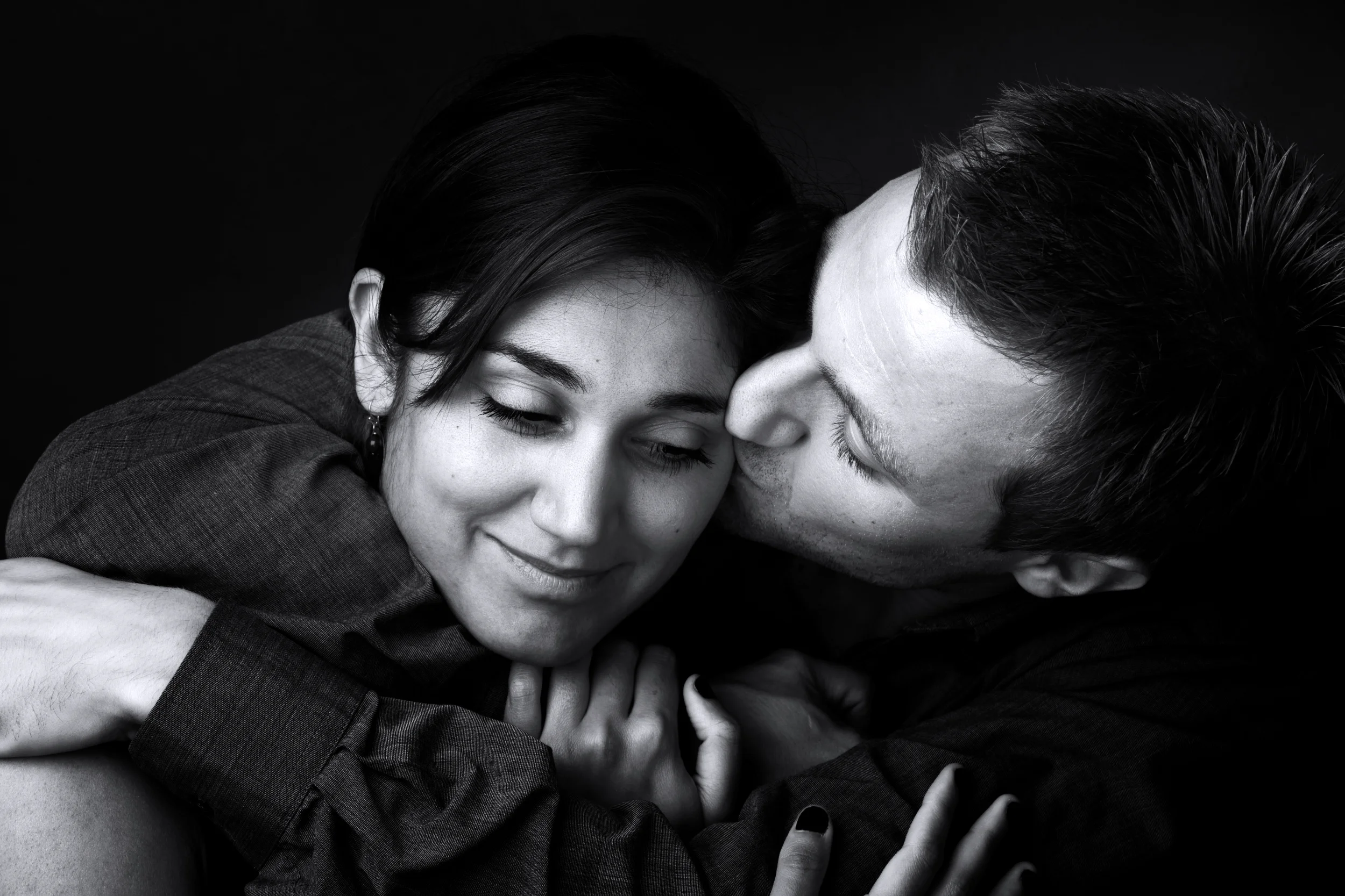 Sort/hvid portrætfoto af mand der kysser kvinde blidt på kinden. Begge er omsluttet af en intim, varm atmosfære.