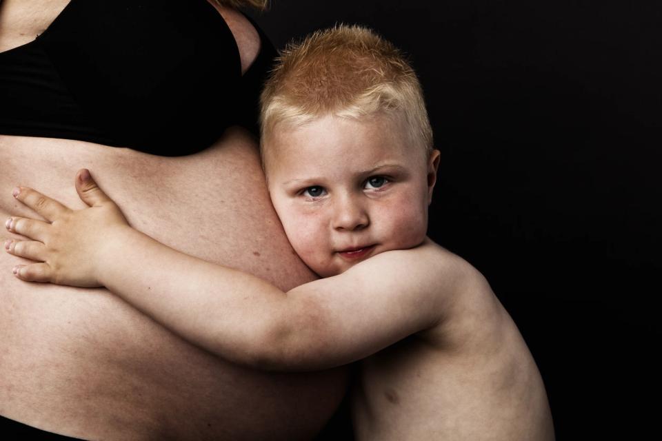 Nærbillede af lyshåret dreng, der omsorgsfuldt krammer sin gravide mors mave mod mørk baggrund, et rørende gravidfoto.