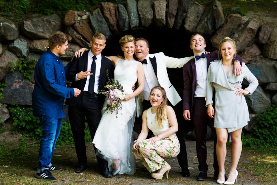 Bryllupsfoto af brudepar med gæster foran en stenvæg. Bruden i hvid kjole, alle smiler opstillet i et uformelt bryllupsbil...