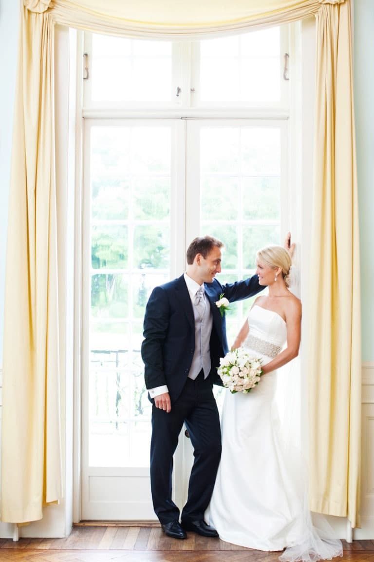 Romantisk bryllupsfoto af brudeparret blidt smilende til hinanden foran et stort vindue på Sølyst. Bruden holder brudebuket.