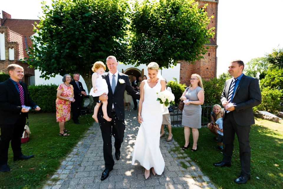 Bryllupsfoto af smilende brudepar der går hånd i hånd, mens gæster kaster ris. Brudgommen bærer datter.