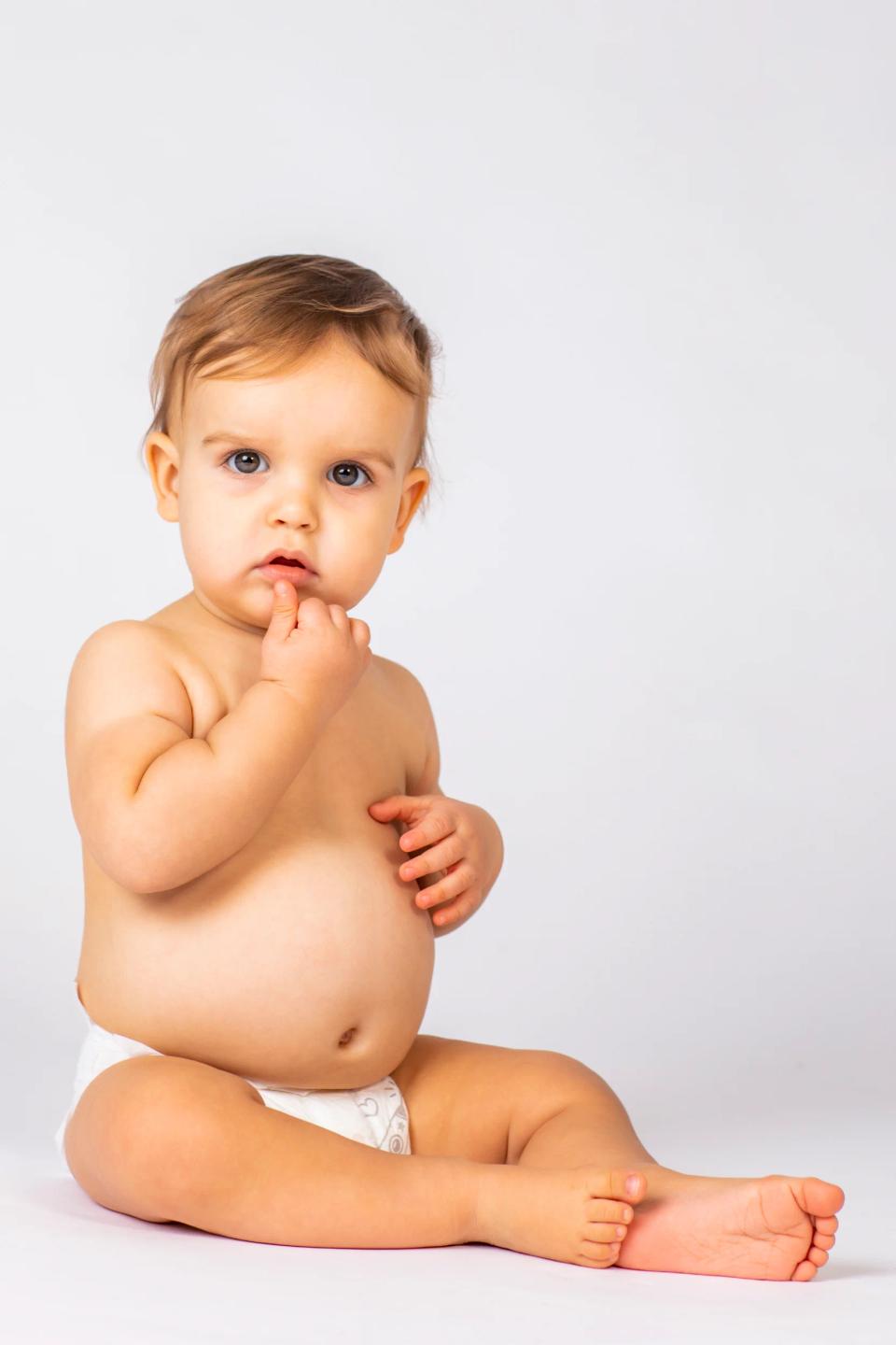 Babyfotografering: Nysgerrig baby med lysebrunt hår sidder i ble og kigger direkte mod kameraet på hvid baggrund.