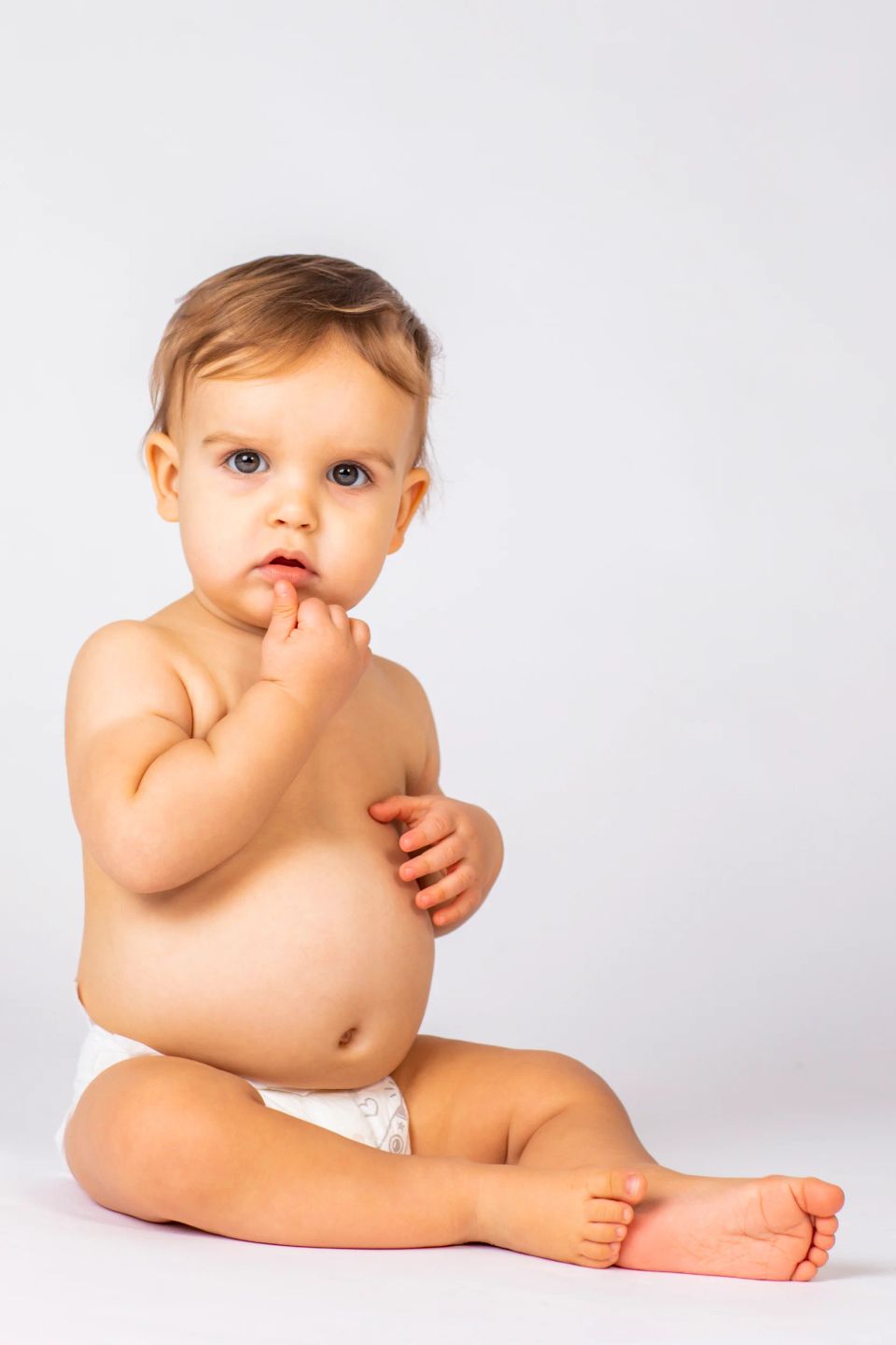 Babyfotografering: Nysgerrig baby med lysebrunt hår sidder i ble og kigger direkte mod kameraet på hvid baggrund.