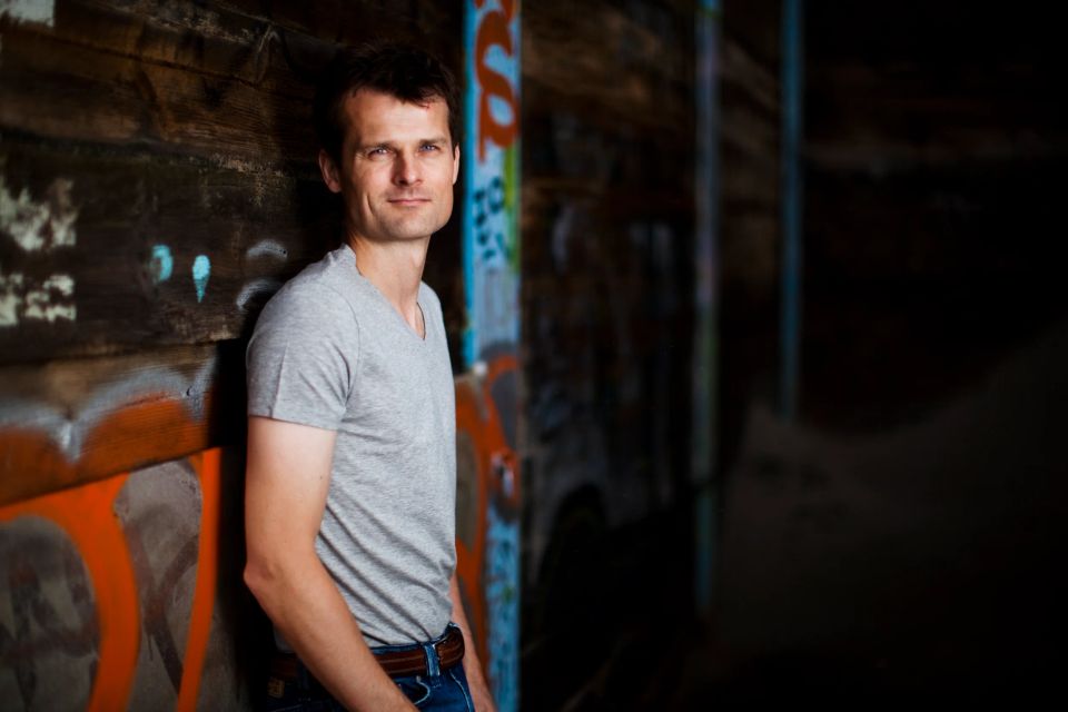 Portrætfoto af smilende mand i grå T-shirt, der læner sig mod en farverig graffitimur med naturligt lys.
