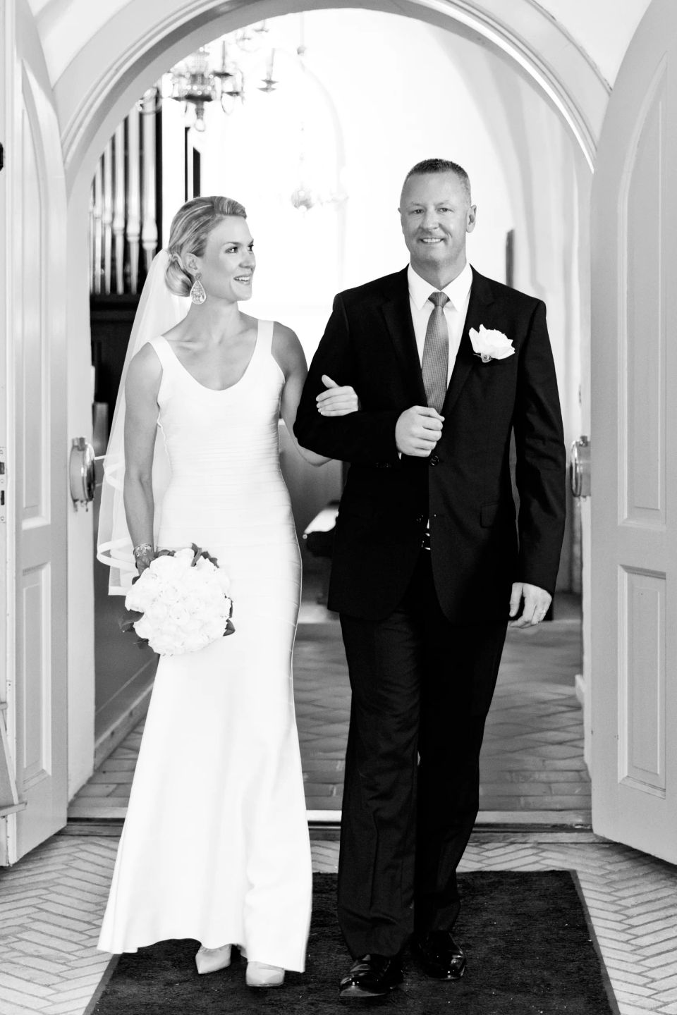 Bryllupsfotografering af brud og gom i sort-hvid. Bruden kigger smilende på gommen, de går ud af kirken i blødt lys.