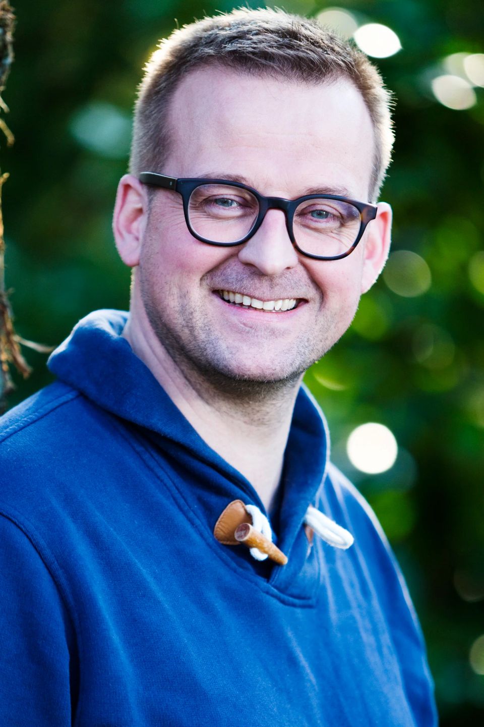 Glad mand med briller i mørkeblå trøje smiler i et portrætfoto med grøn sløret baggrund og naturligt lys.
