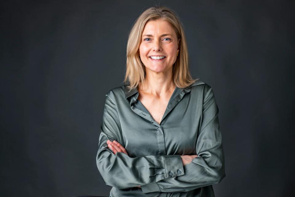 Portrætfoto af smilende kvinde med blondt hår og blå øjne i grøn silkeskjorte, hænderne foldet, foran mørk baggrund.