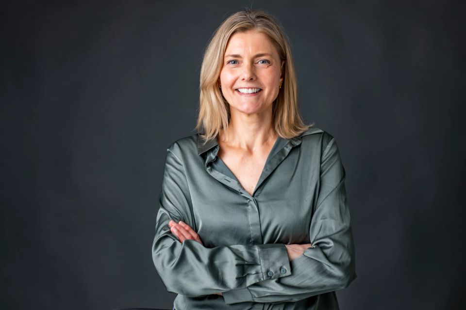 Portrætfoto af smilende kvinde med blondt hår og blå øjne i grøn silkeskjorte, hænderne foldet, foran mørk baggrund.