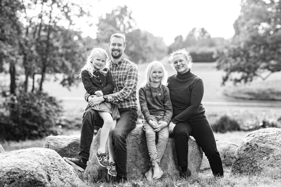 Familiefotografering: Smilende familie på fire sidder på sten udendørs med baggrund af træer i varmt modlys.