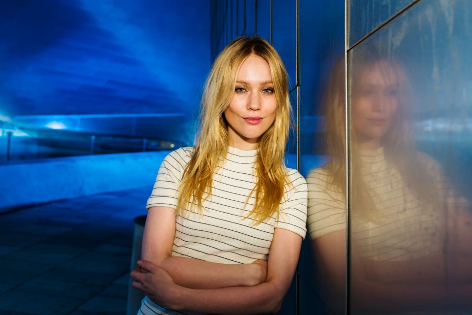 Autentisk portrætfoto af kvinde med lyst hår, der smiler skævt, lænende mod en mørkblå væg med refleksion.