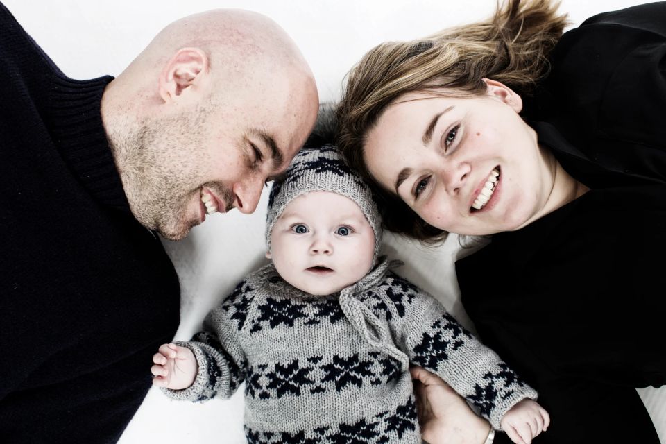 Kært babyportræt: Glad mor og far smiler til deres nysgerrige baby i striktrøje og hue, der kigger direkte ind i kameraet.