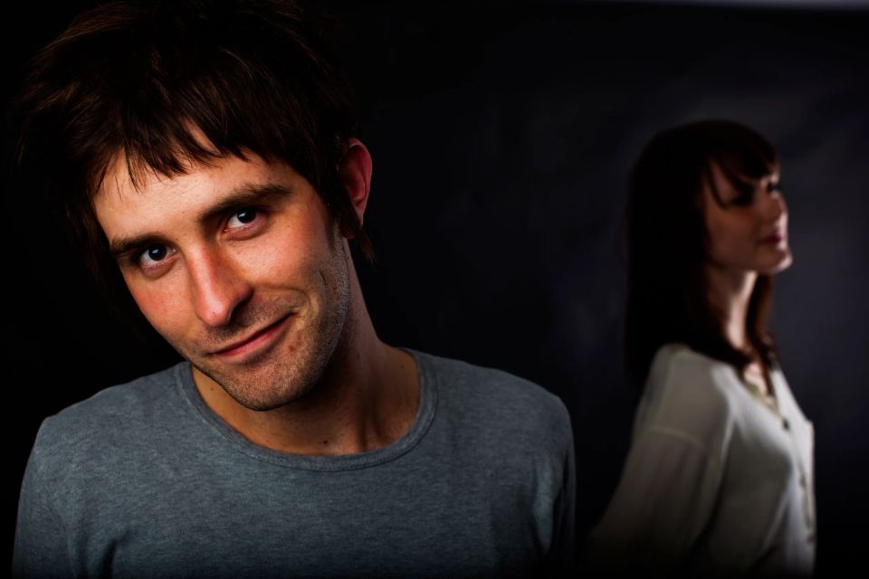 Portrætfoto; smilende mand med mørkt hår, skægstubbe, grå t-shirt i skarpt lys, kvinde i baggrunden i skygge.