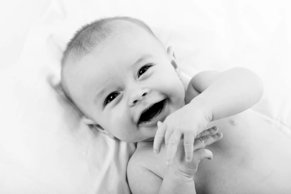Glad babyfotografering i sort-hvid af en smilende baby, der ligger på ryggen med åben mund og arme løftet.