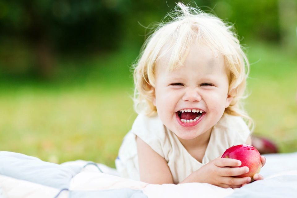 Glad pige med lyst hår, der griner og holder et rødt æble, liggende på tæppe i haven. Børnefotografering med leg og glæde.