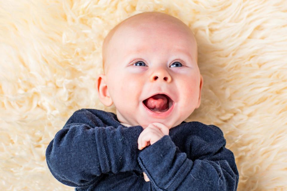 Sødt baby/nyfødt portrætfoto af glad baby i mørk trøje med åben mund, liggende på blødt lyst tæppe i naturligt lys.