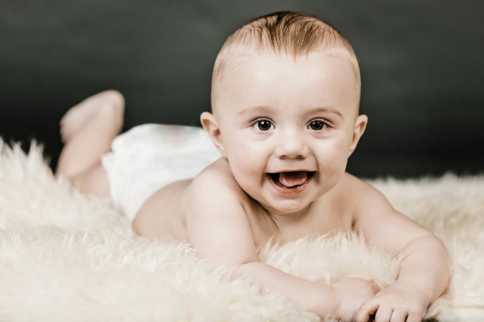 Glad baby ligger på maven på et hvidt pelstæppe med mørk baggrund, giver et stort smil til kameraet.