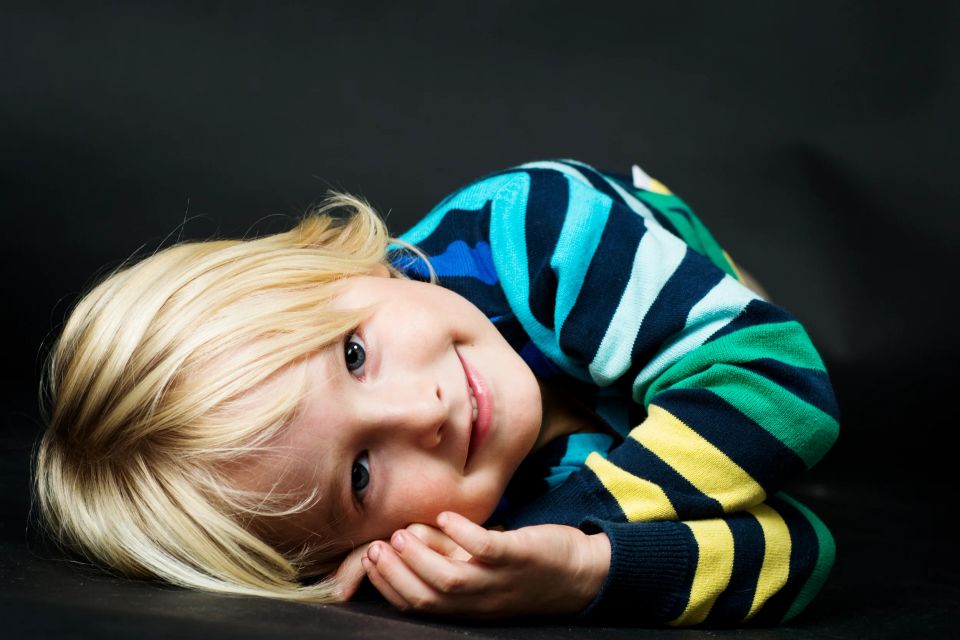 Børneportræt af smilende lyshåret barn med ansigtet vendt mod beskueren i farverig stribet trøje liggende på siden.