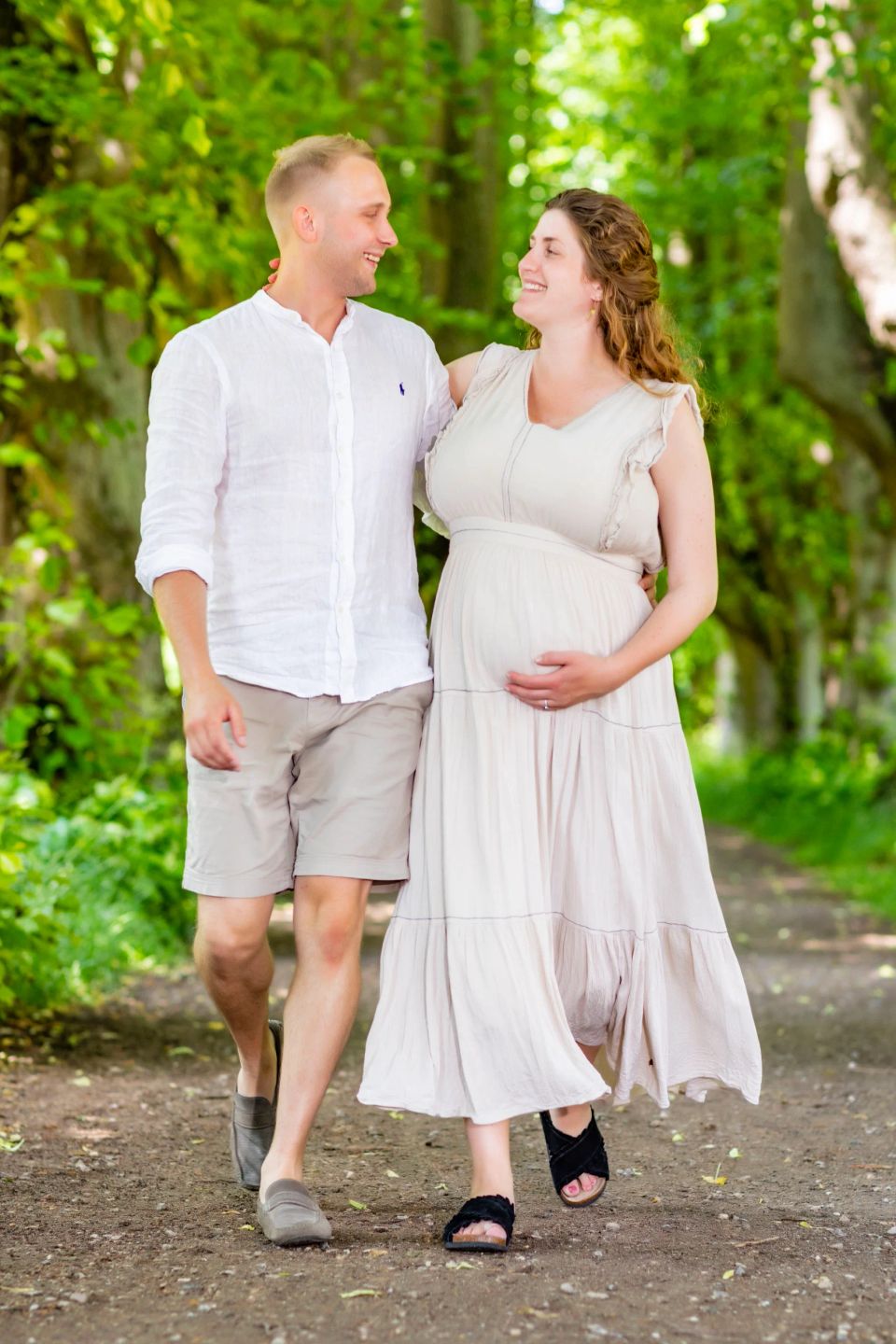 Gravid fotografering: Parret går hånd i hånd og smiler kærligt til hinanden på en grøn skovsti i blødt sollys.