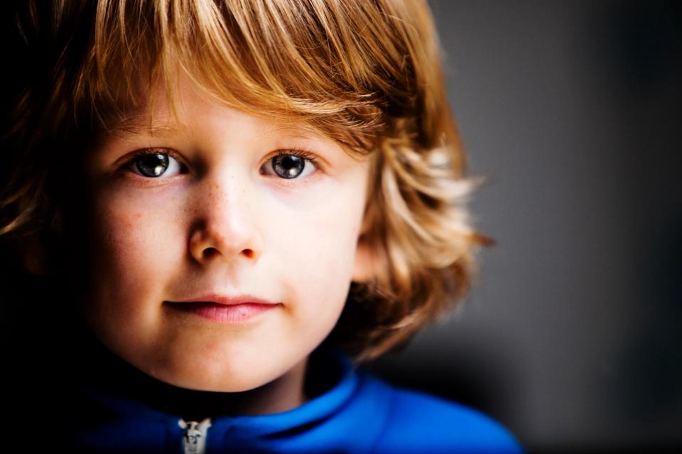 Nærbillede af et barn med lyst hår og udtryksfulde øjne, iført en blå bluse, i blødt sidelys. Portrætfoto.